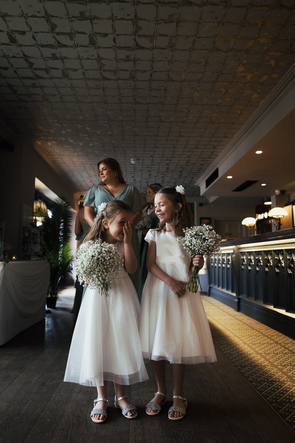 Flower girls in ivory tulle bridesmaid dresses from Monsoon giggle while waiting in the bar for the wedding ceremony at The Lion House, Chelmsford, Essex (CM3 3JA). A bridesmaid in a Mori Lee chiffon dress (style #21667) with flutter sleeves and a front slit stands nearby, creating a joyful and candid wedding moment.