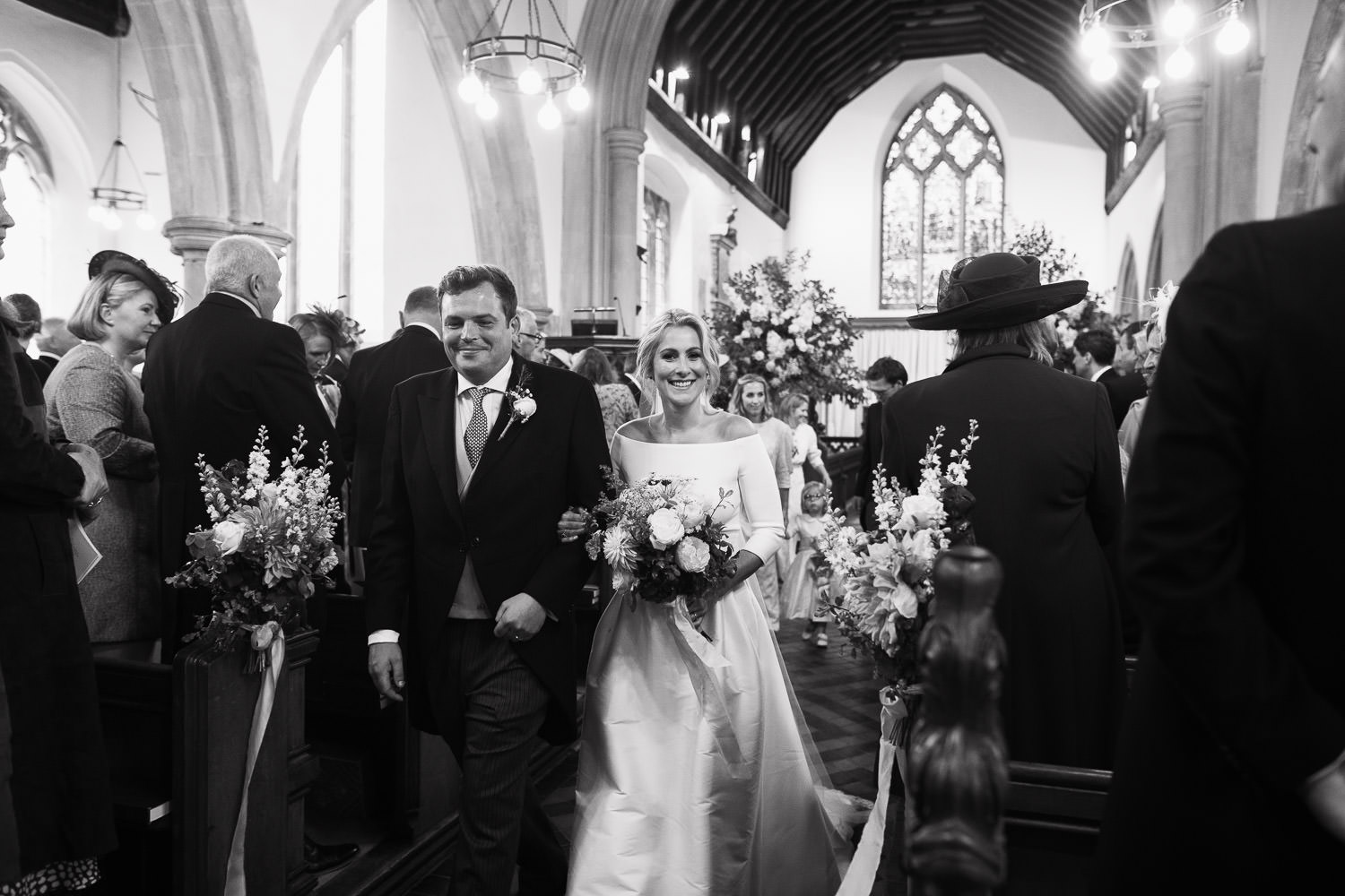 Newly married couple walking down the aisle after their ceremony at the Church of St John Baptist in Danbury, Essex. Guests stand in the pews, celebrating the moment. Dress: Miss Bush. 55 Main Rd, Danbury, Chelmsford, CM3 4NG.