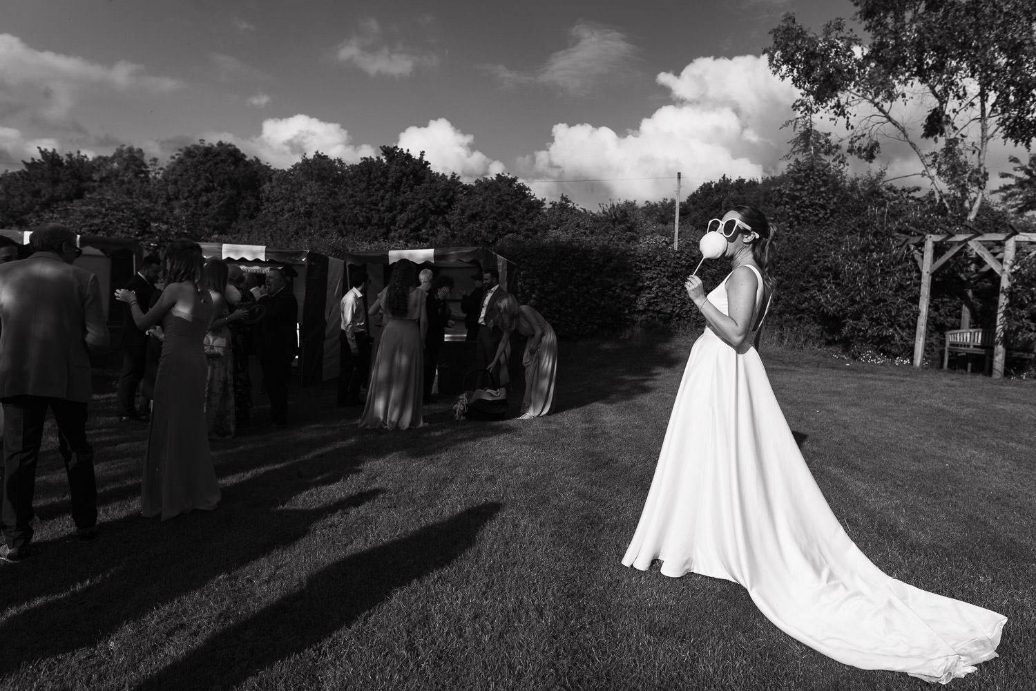 Alpheton Hall Barns Wedding Photography: Bride in a white satin dress and oversized sunglasses enjoys candy floss in the sun. Guests have fun playing fairground games.