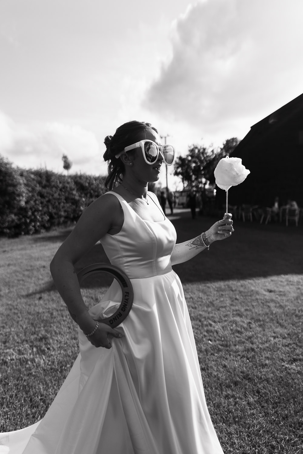 sunglasses holds a stick of candy floss and a frisbee, in the bright sunshine outside Alpheton Hall Barns in the stackyard wedding garden. By Essex Wedding Photographer Tracy Morter.