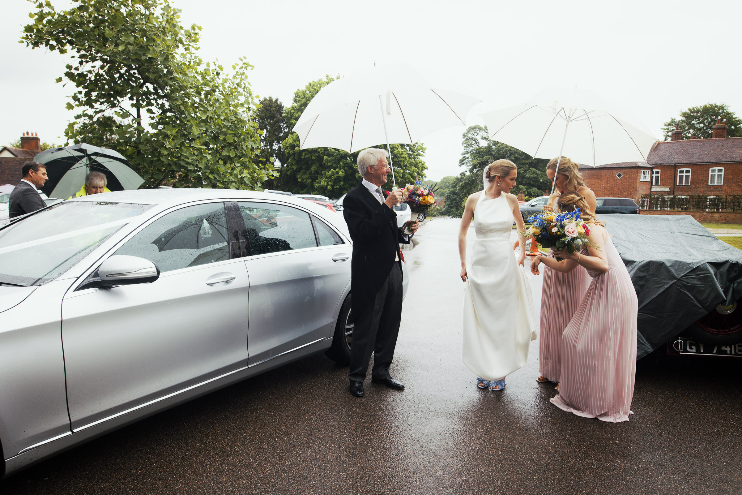 Bride in a Jesús Peiró 260 dress from Miss Bush stands outside a church on a rainy day in Danbury, Essex CM3 4NG, as her dad and bridesmaid hold an umbrella. Captured by a documentary-style Essex wedding photographer from South Woodham Ferrers.