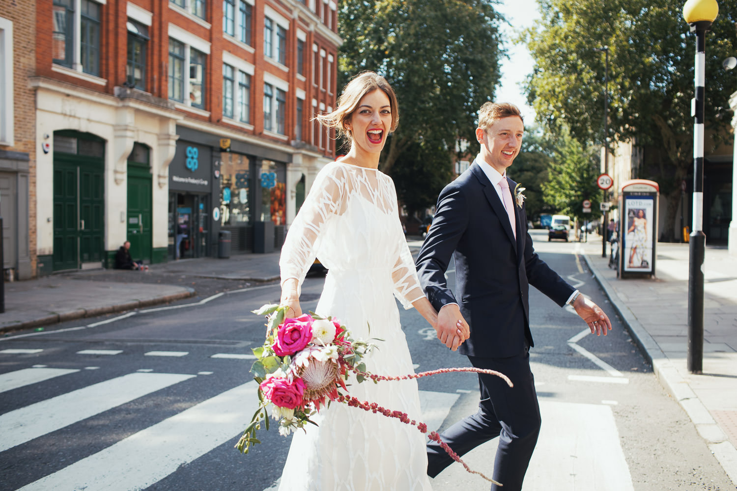 The bride and groom cross Tysoe Street near Exmouth Market. The bride is wearing a Halfpenny dress and holding pink flowers, smiling widely at the camera. The groom is dressed in a dark suit with a pale pink tie. In the background, the Co-op and a phone box are visible. A wedding photojournalism-style photograph taken in London.