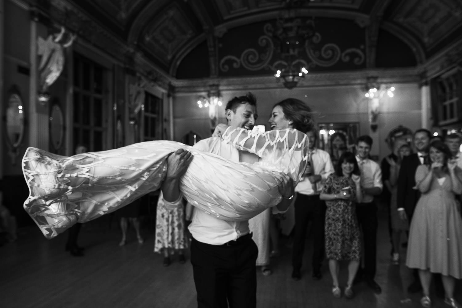 Groom picks up his wife and spins her around at a wedding reception at the Old Finsbury Town Hall. The bride, wearing the Willow dress from Halfpenny London. Captured in a documentary wedding photography style.