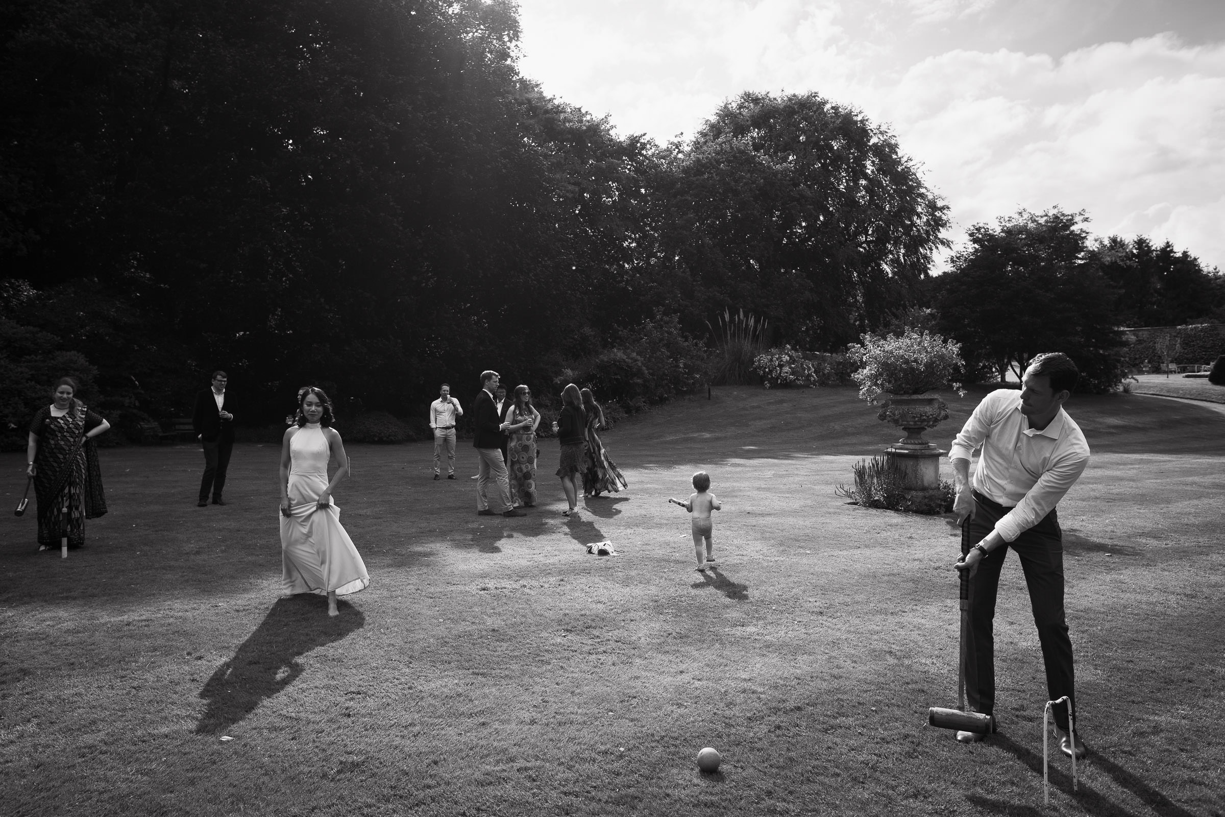 Man holding a croquet mallet, a barefoot bridesmaid lifting her dress as she walks across the lawn, and a toddler wandering off while wedding guests chat on the main lawn of Homme House.