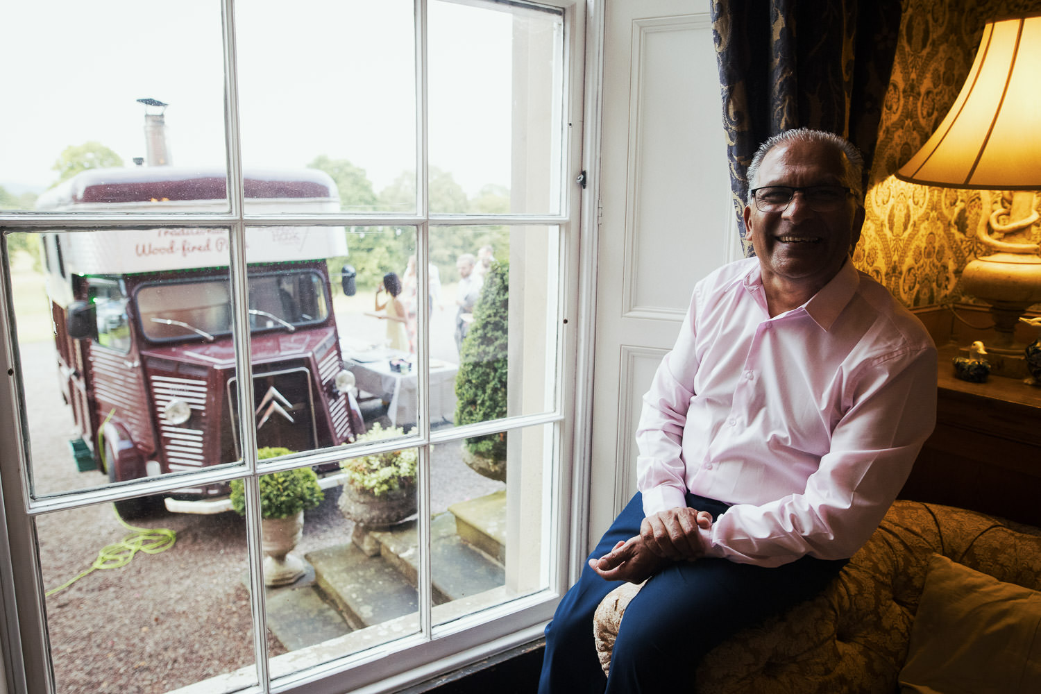 An unposed portrait photograph of a man, the father of the groom, sat on a sofa at the wedding venue Homme House. A pizza van can be seen outside the window.