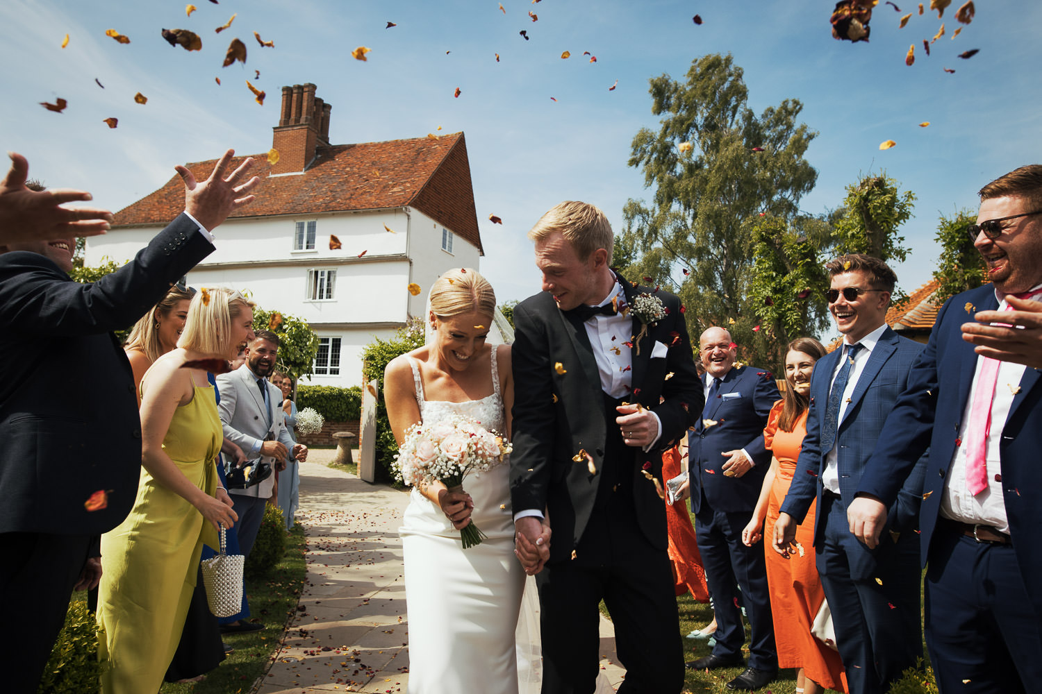 Newlyweds walking through confetti tunnel at Houchins Essex. Groom in black tie suit from The Groom’s Room, Colchester. Bride in Jack dress from Halo + Wren. Documentary wedding photography.