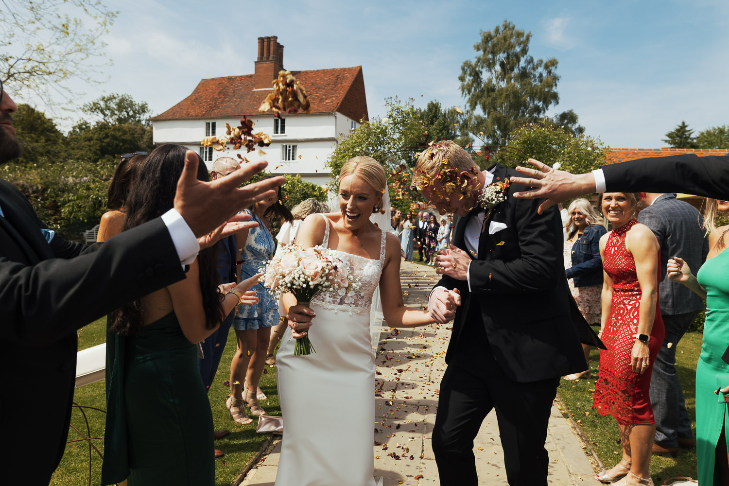 Newlyweds walking through confetti tunnel at Houchins Essex. Groom in black tie suit from The Groom’s Room, Colchester. Bride in Jack dress from Halo + Wren. Documentary wedding photography.