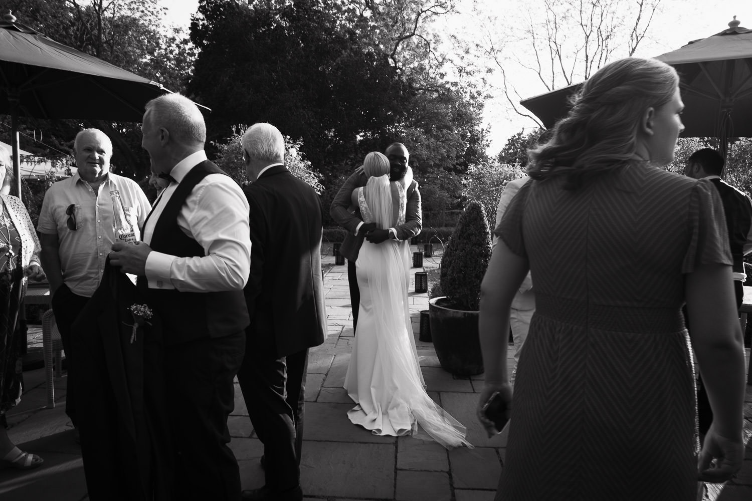 Documentary wedding photography by Tracy Morter at Houchins, an Essex wedding venue. The bride, wearing the 'Jack' dress by Made With Love Bridal from Halo + Wren bridal shop, hugs a wedding guest on the terrace in the evening. CO6 1RT