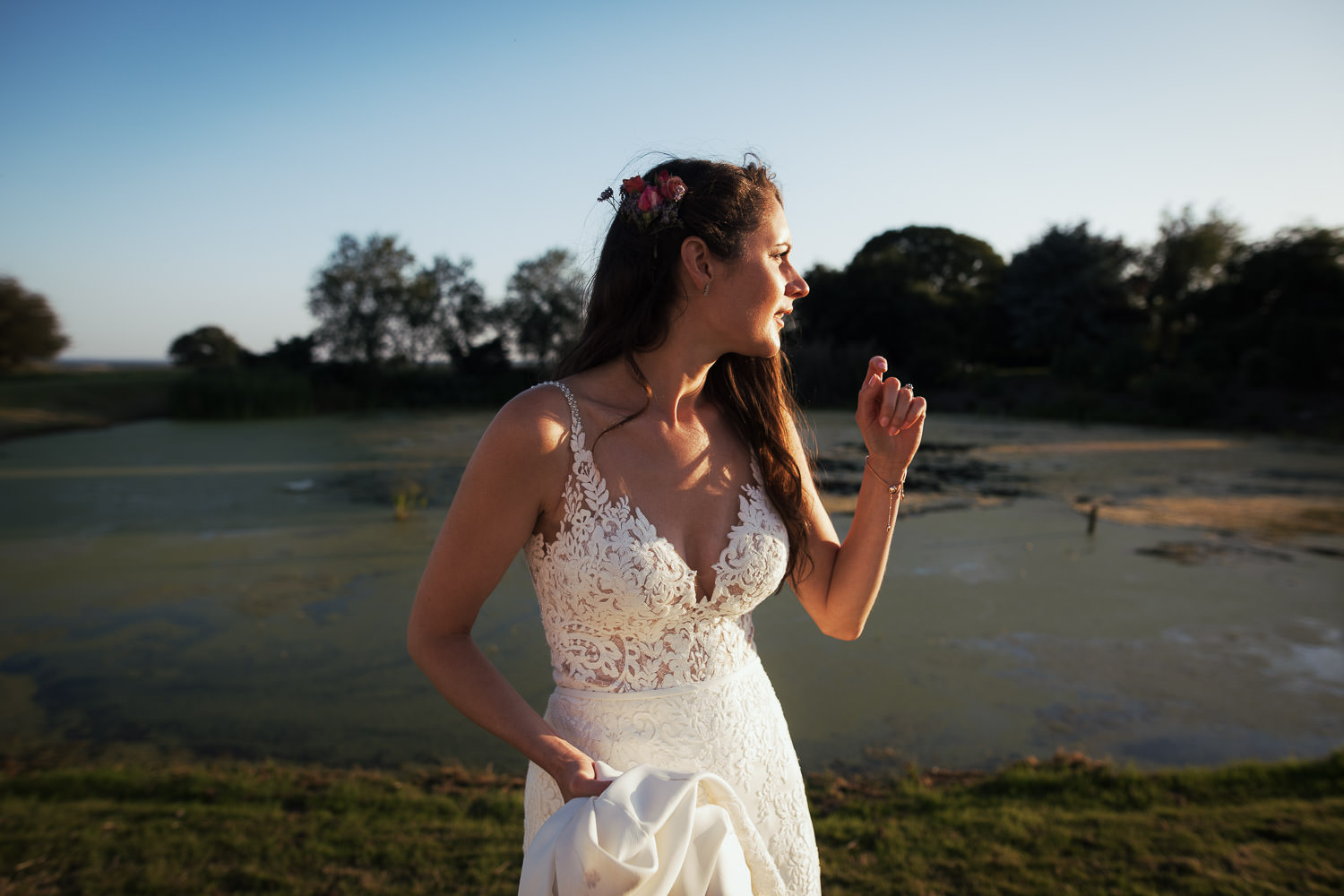 The bride in a lace wedding dress stood by a pond. The sun is setting so the light is low and casting shadows. The wedding is in Essex near South Woodham Ferrers. Candid wedding photographer from Essex. Bride is wearing Stella York 6648 from The Wedding Shop in Colchester.