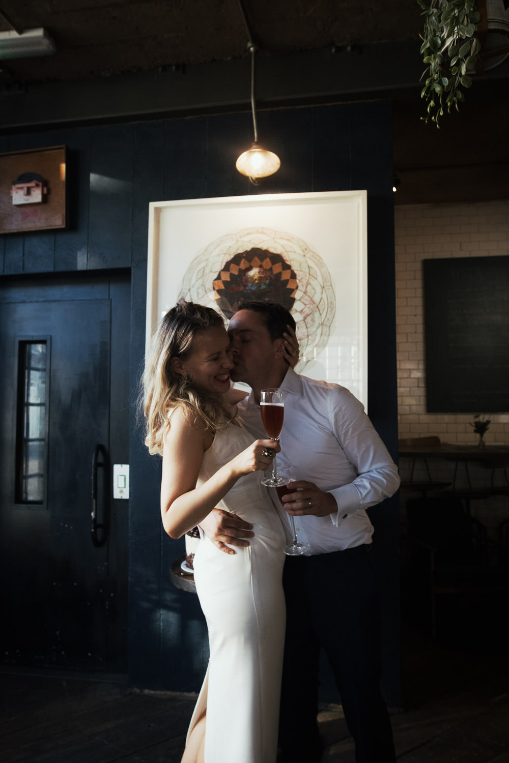 Essex Documentary Wedding Photographer: Candid Kiss at Wedding Reception A newly married couple at their wedding reception at Never For Ever restaurant. London wedding. They are holding full champagne flutes.