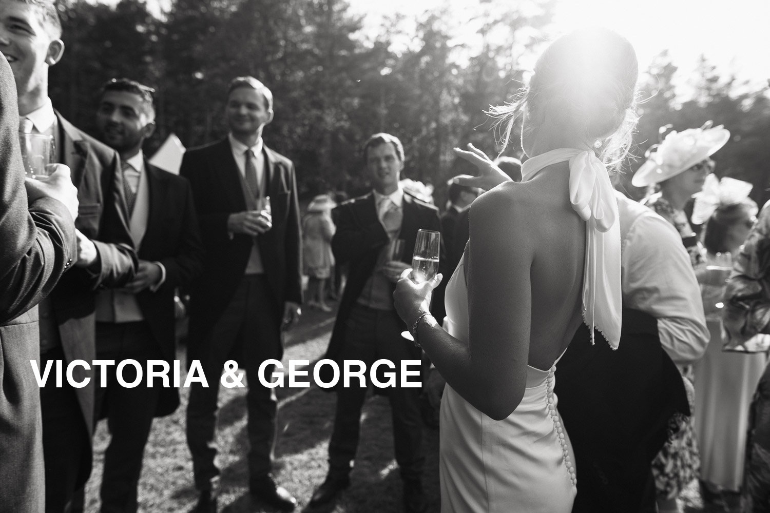 View from behind a bride on her wedding day, drinks in the sun. She's holding glass of champagne, men in the background wearing suits. Candid Norfolk Wedding photography. Katrine Mogensen bespoke bridal gown.