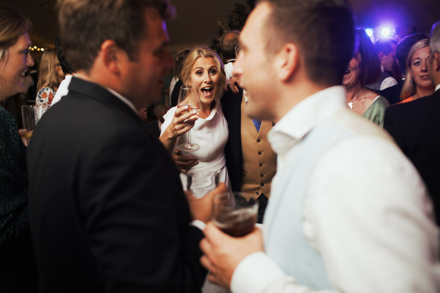 Bride holding a glass of wine, surrounded by guests on the dance floor in a Sail & Peg marquee at a vineyard near South Woodham Ferrers. Essex wedding photography, wearing a Miss Bush Bridal dress