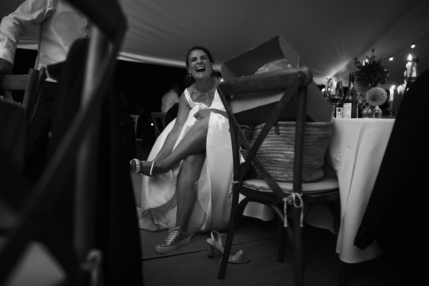 Documentary-style wedding photograph of a bride in a white wedding dress sitting at a table inside a marquee by County Marquees Ltd, changing her shoes and laughing. Captured at a wedding in Thaxted, Essex. Makeup by Sarah Drummond