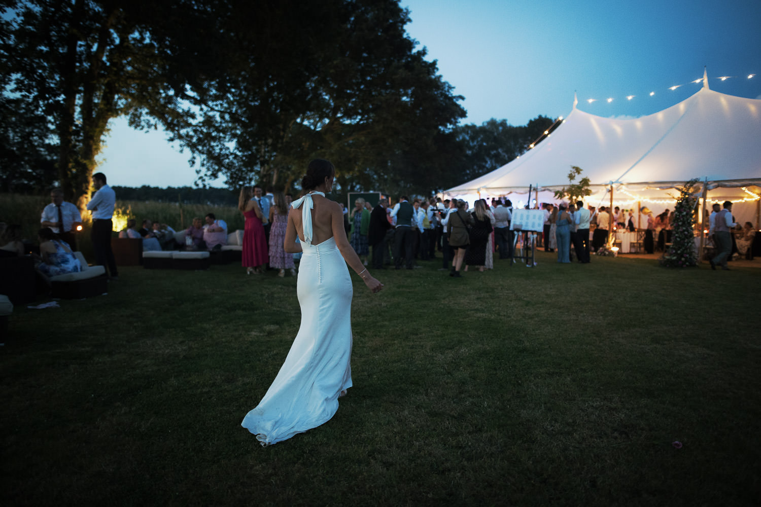 A candid evening photography of a marquee wedding in Norfolk. Morton's Event Hire. Trees in the background. Bride seen from behind walking towards the marquee. Wearing a halter neck dress by Katrine Mogensen Bridal Couture.