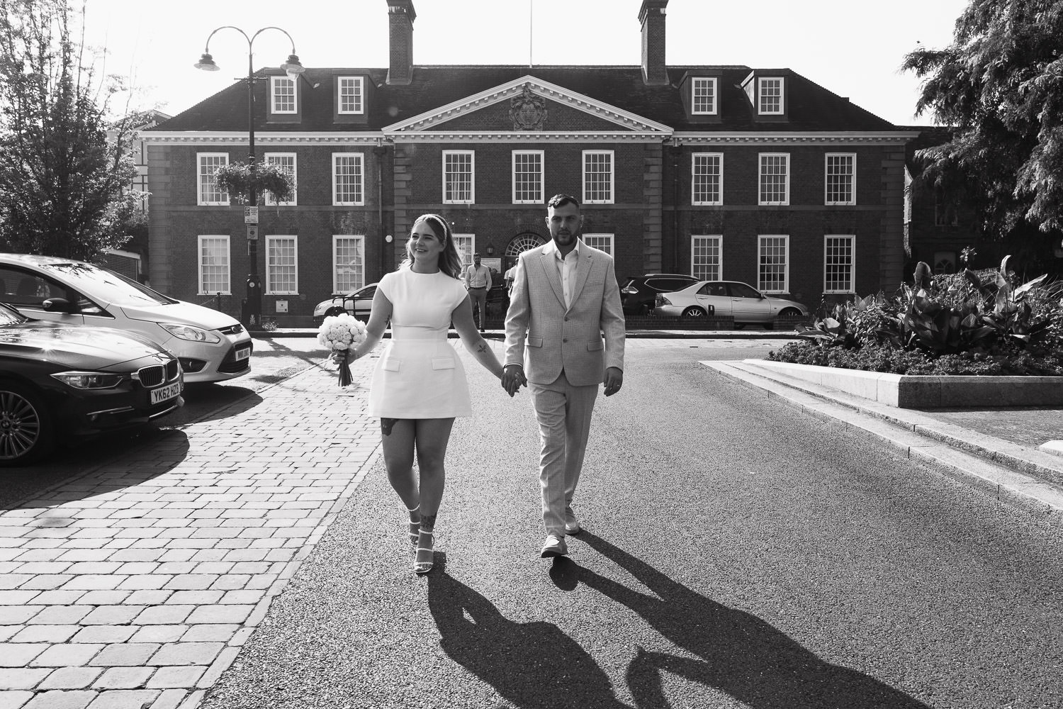 Couple leaving the Bury St Edmunds Register Office Suffolk wedding ceremony in the sunshine. IP33 1UZ Bride is wearing the Odd Muse The Ultimate Muse Cap Sleeve Mini Dress in White. Groom is wearing a Moss slim fit off white check suit.