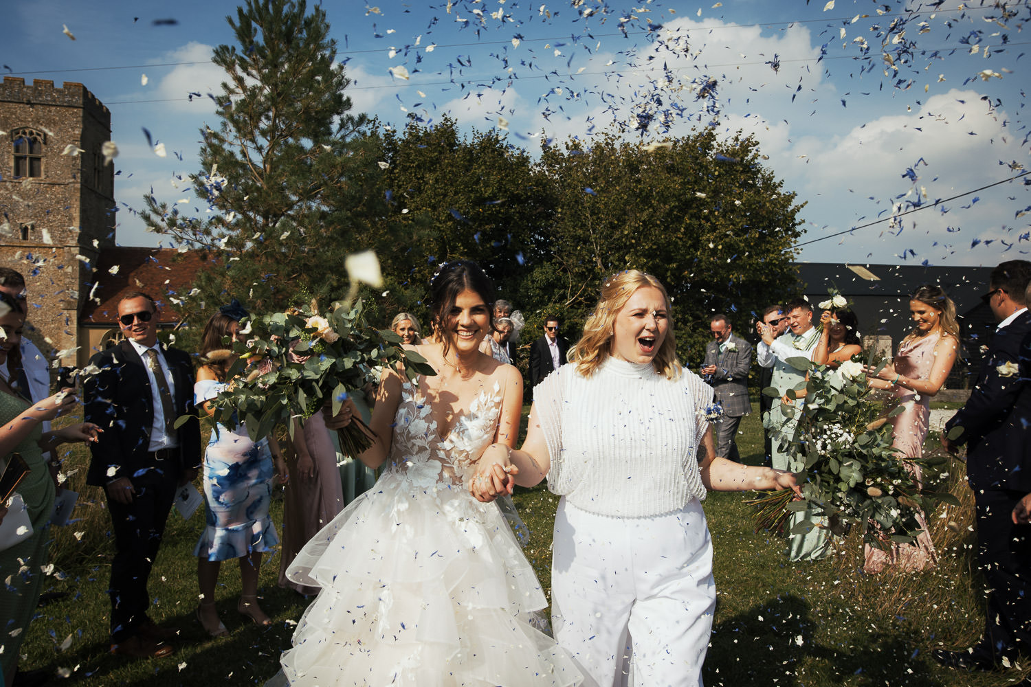 A confetti tunnel for the two brides at Alpheton Hall Barns in the sun. Guests throw blue and white confetti. Brunette bride is wearing a dress called Montgomery by WTOO by Watters Montgomery. She's holding hands with her wife who is wearing a white Joanna Hope jumpsuit. Both holding bouquets by Suffolk florist Wild + Mae.