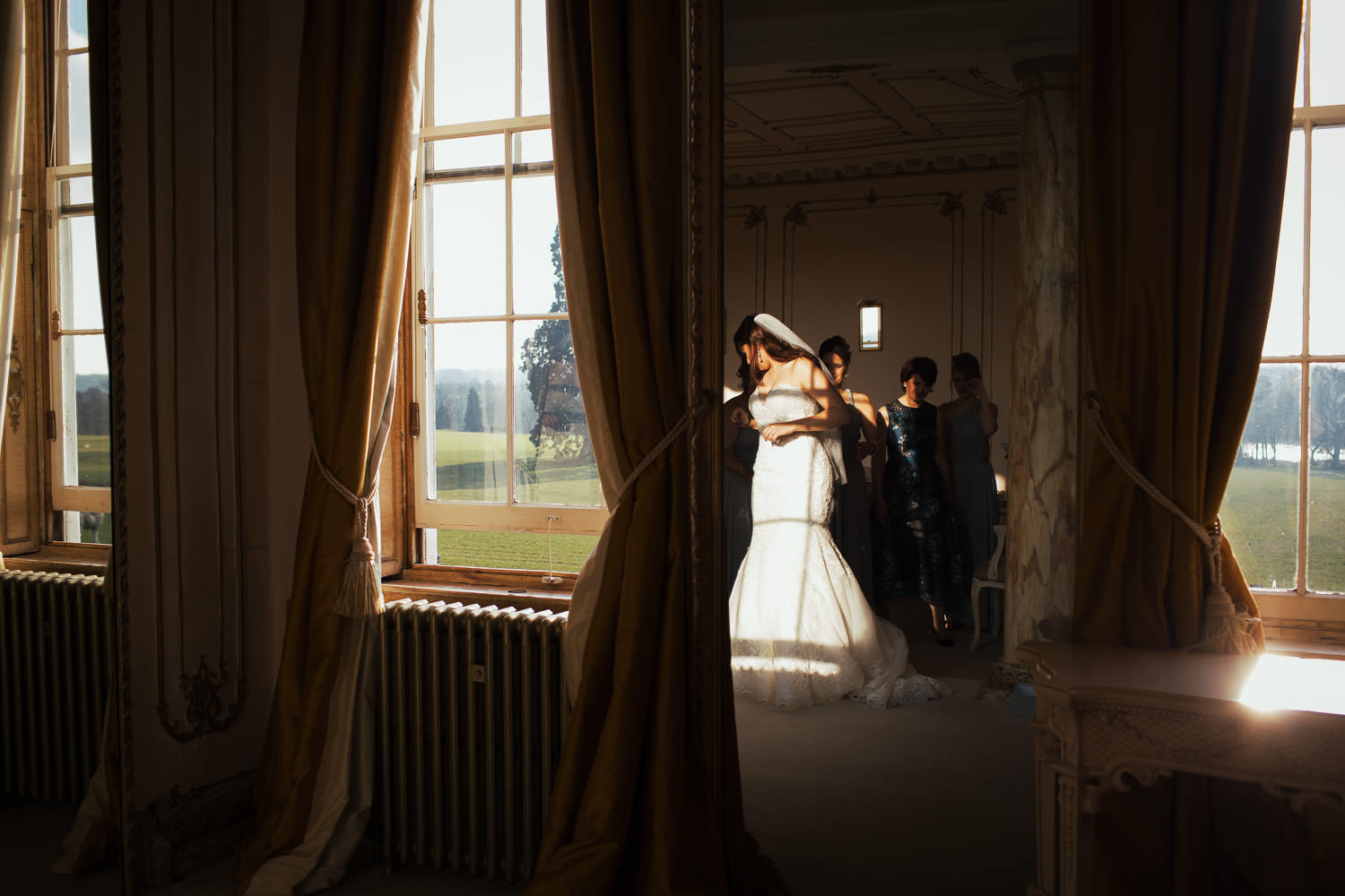 In the Rococo suite at Gosfield Hall. A reflection in a mirror of a bride in a fishtail wedding dress. Style 2713 by Mori Lee Madeline Gardner. The shadow of the window frame lands on her dress. Outside the windows you can see grass and trees. It's a sunny winter day.