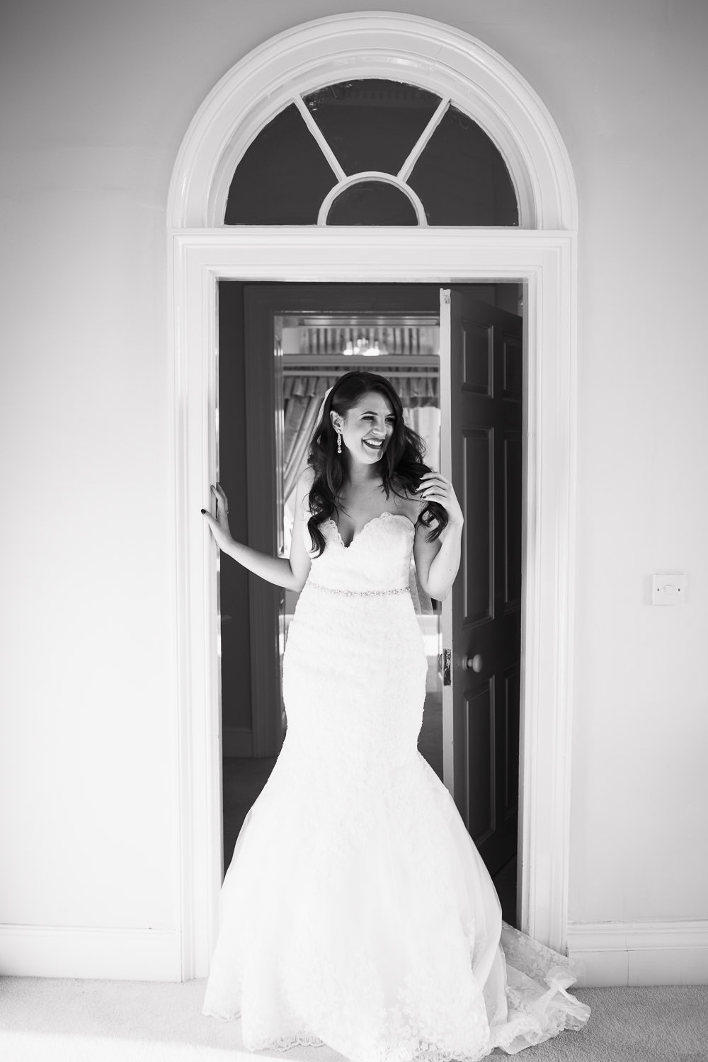 Gosfield Hall Wedding: Bride in a lace mermaid style wedding dress, style 2713 by Mori Lee. She has long dark hair and red lipstick. She is smiling and standing in the doorway of the Queen's Apartment. An unposed wedding photograph.