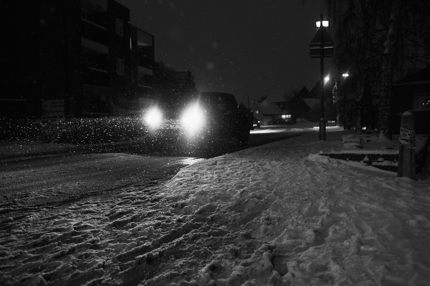 A car approaching the South Woodham Ferrers station on Hullbridge Road at night, amidst a snowy scene. In the background, The Cedars flats are visible.