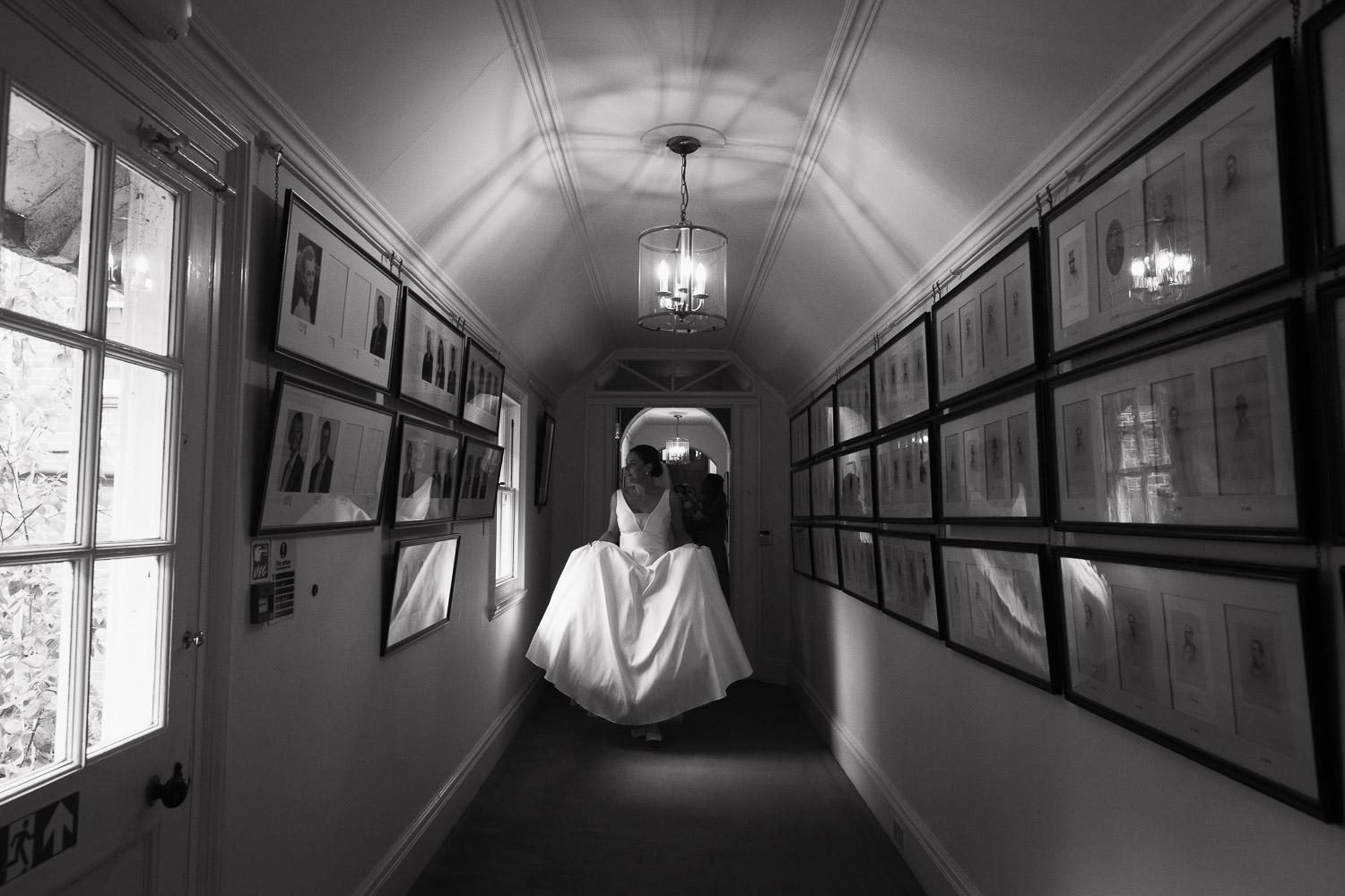 At the Jockey Club Rooms in Newmarket the bride walks down a corridor holding up her wedding dress. Rosa Clará wedding dress with deep-plunge V-neckline and pockets. from BOA Boutique in Surrey. Pictures on the wall, the glass reflecting the bride. Documentary wedding photography near Cambridge.