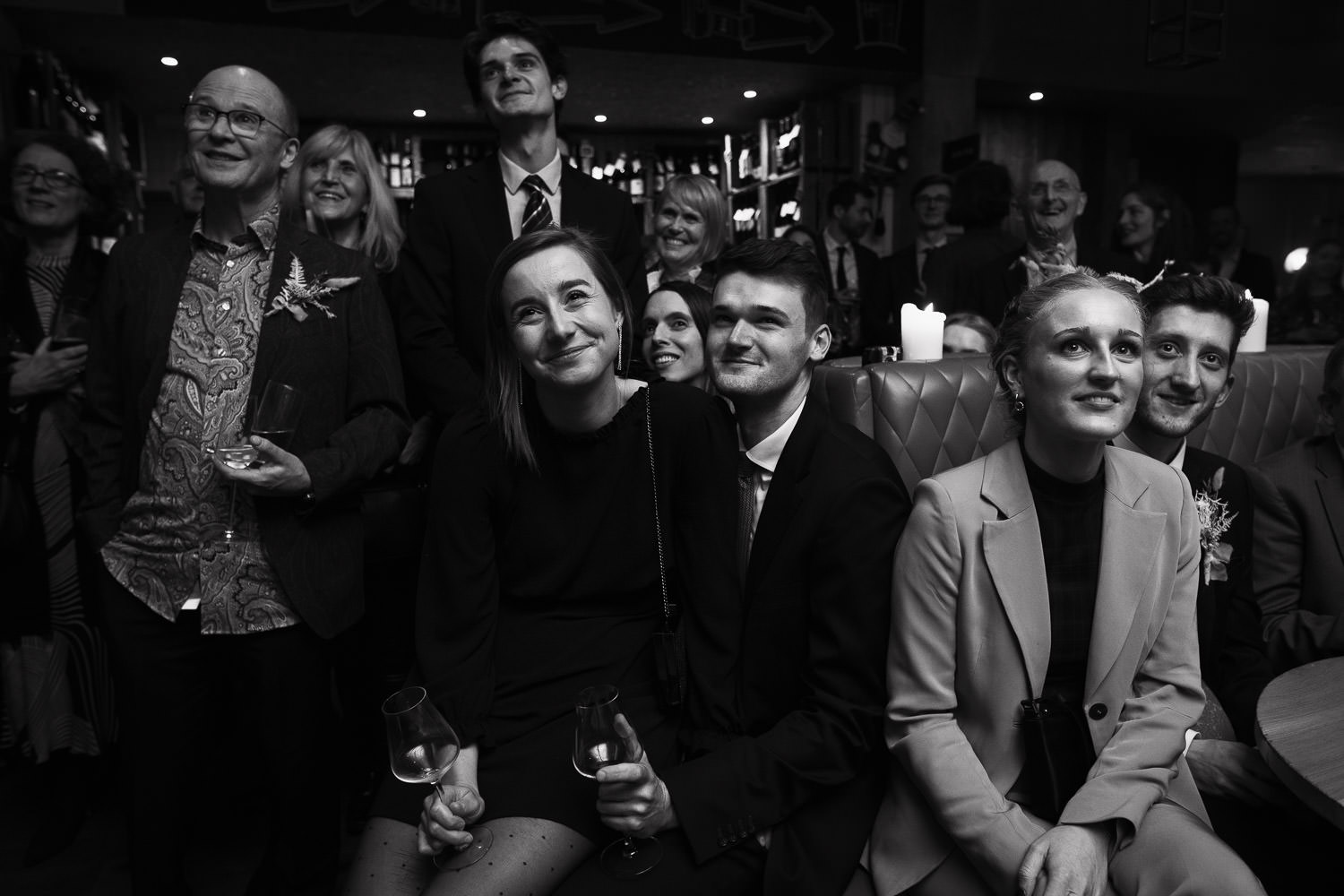 Humble Grape London Wedding Reception: Smiling woman in dotty tights sits on man's lap. Surrounded by guests, they are watching speeches off camera.