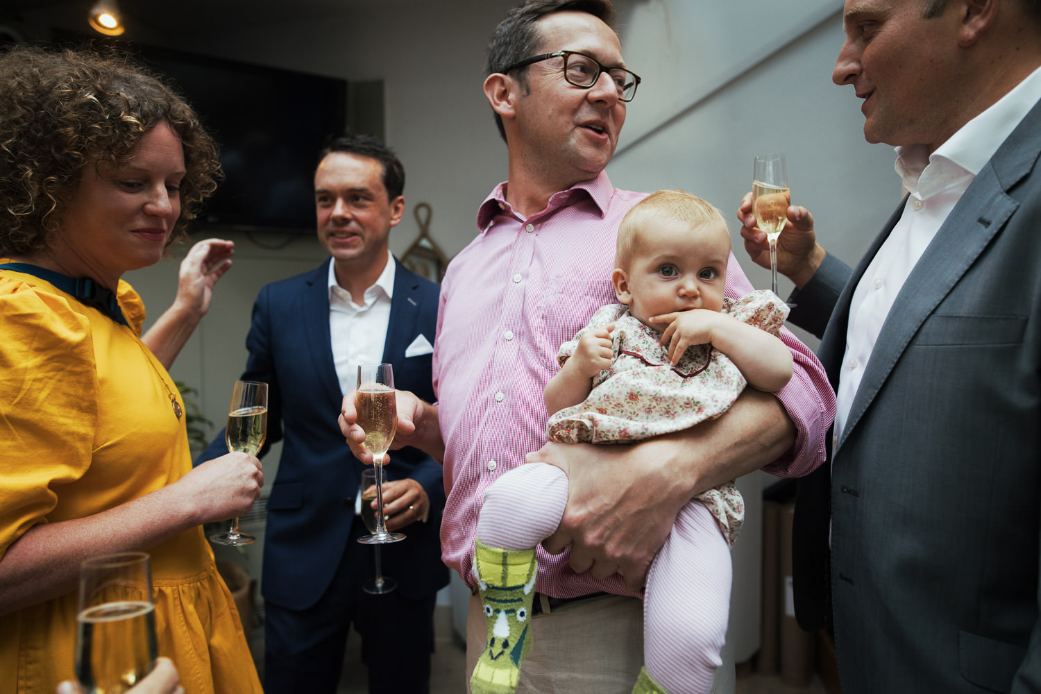 Man in a pink shirt holding a glass of wine and a baby. The baby is breaking the fourth wall and looking at the camera.