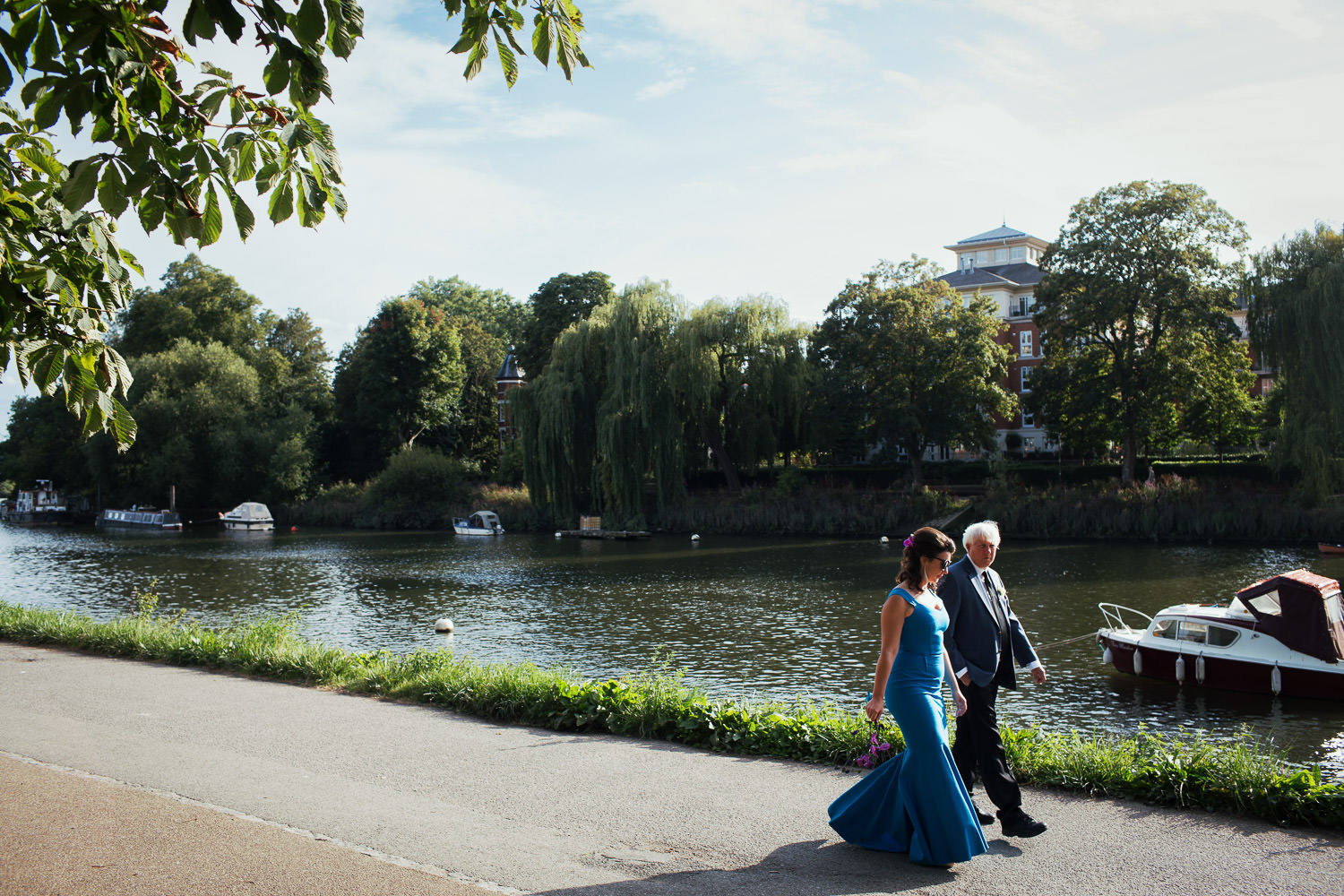 Bride wearing a blue dress by Roland Mouret walks along the Richmond River Thames Towpath with her dad. The willow trees hang down.