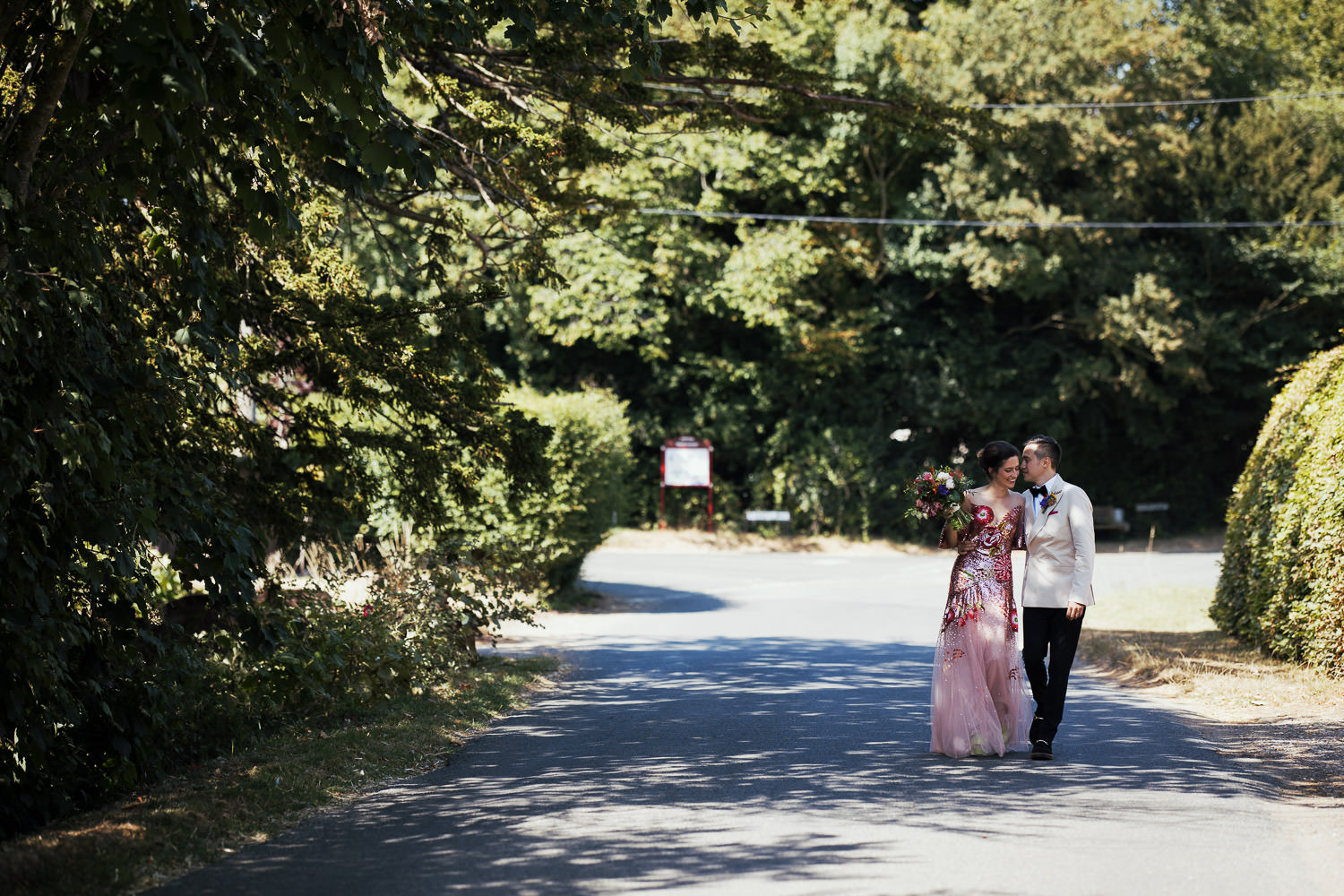 A bride and groom walk down the road in Lawshall after their wedding ceremony. The bride is wearing a Temperley carnation sequin gown, the groom is wearing black tie with a white dinner jacket.