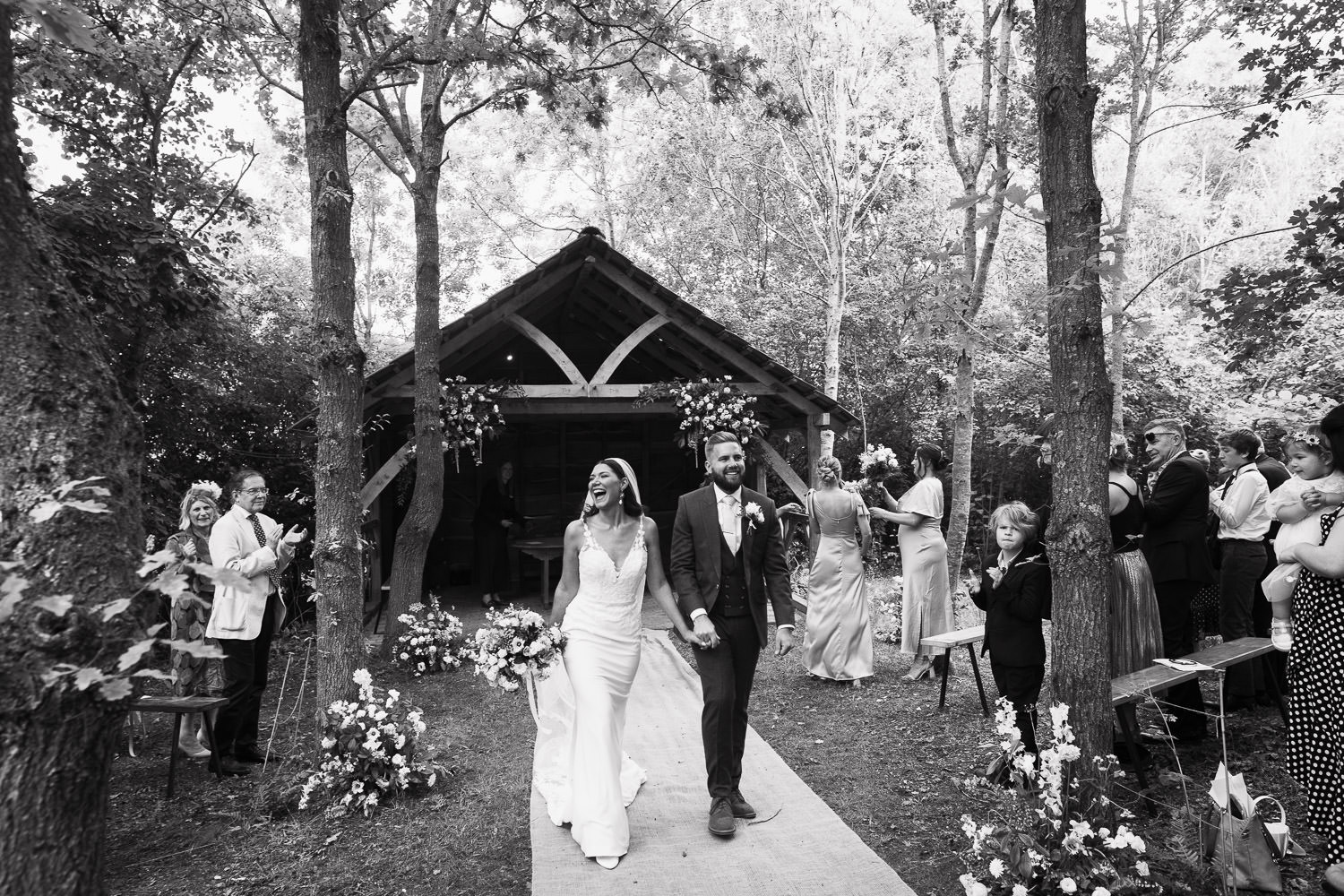 Newly married couple walking down the woodland ceremony glade aisle at Alpheton Hall Barns in Suffolk. Bride is wearing Maggie Sottero style dress 22MW548B01