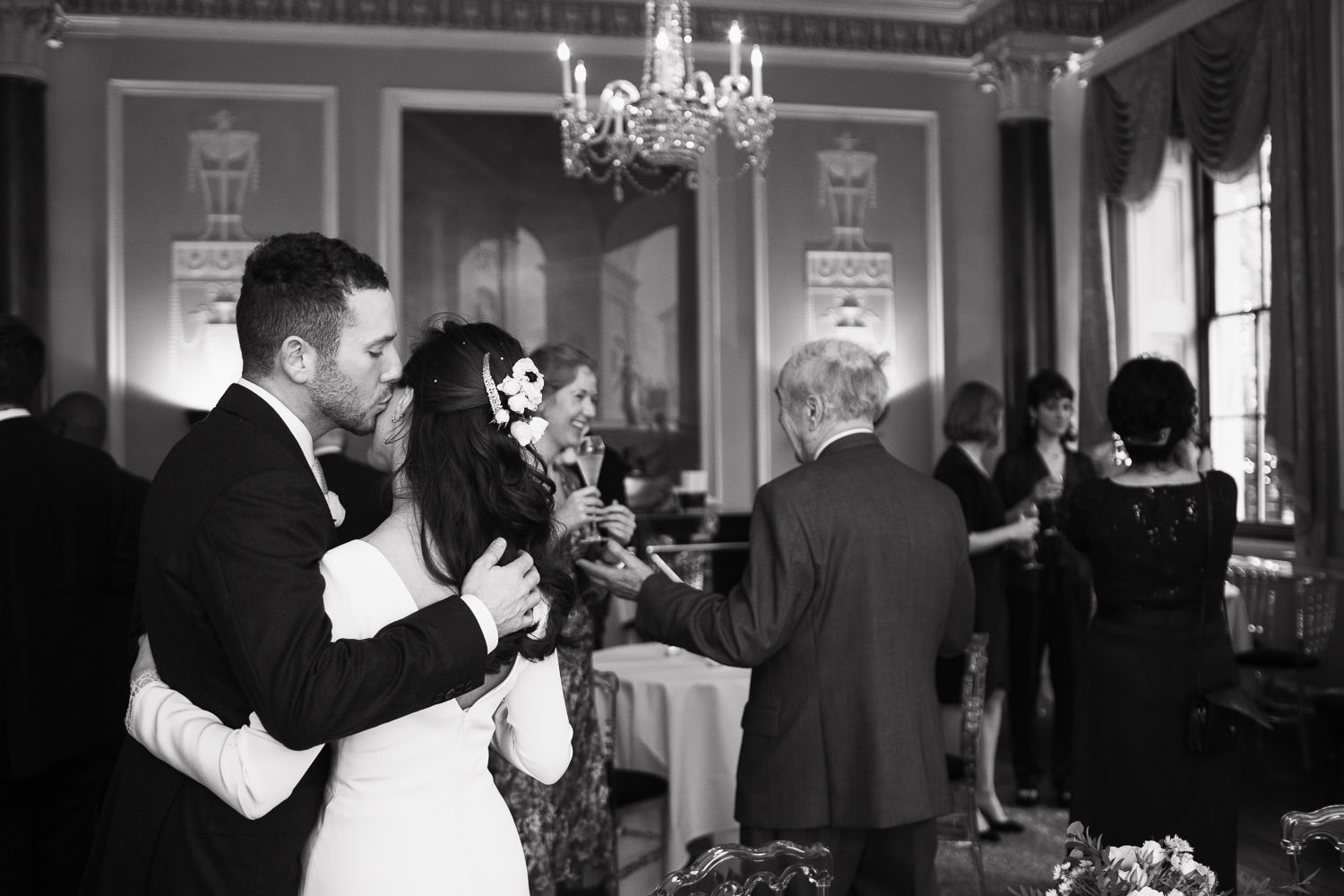 In the front parlour of the Home House a man puts his arm around a woman and kisses her on the cheek. They just got married. There are wedding guests in the background having drinks.