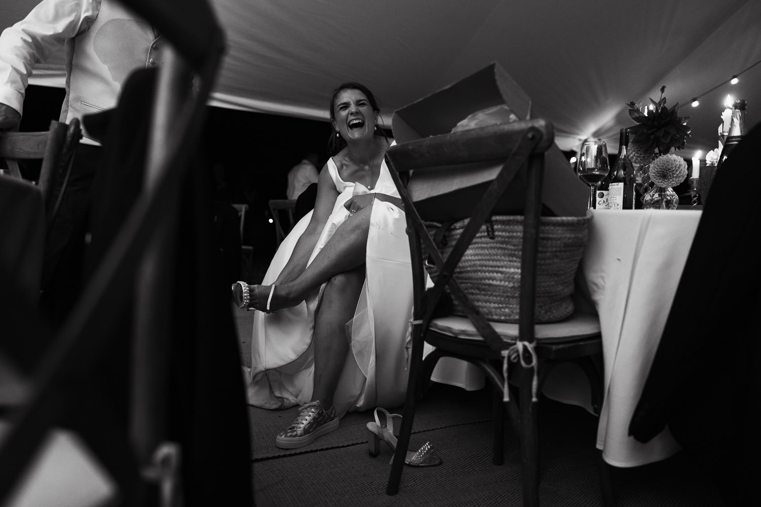 Documentary-style wedding photograph of a bride in a white wedding dress sitting at a table inside a marquee by County Marquees Ltd, changing her shoes and laughing. Captured at a wedding in Thaxted, Essex. Makeup by Sarah Drummond