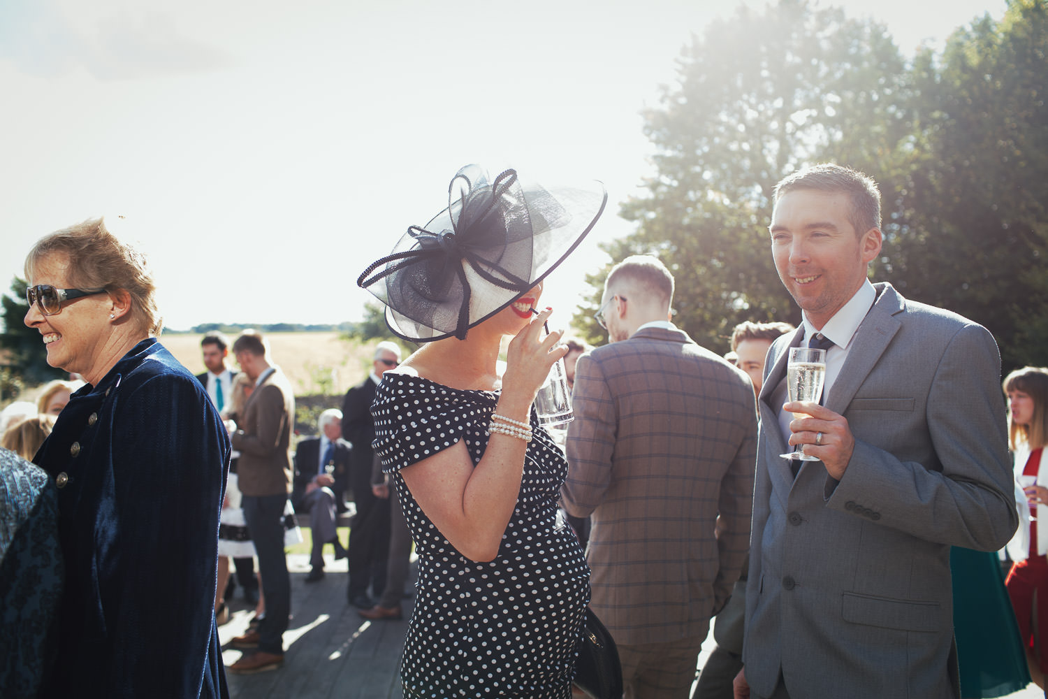 Pregnant woman in a big hat drinking in the sunshine. A wedding at a barn in the Cotswolds. Fosscross Ln, Cirencester GL7 5BA