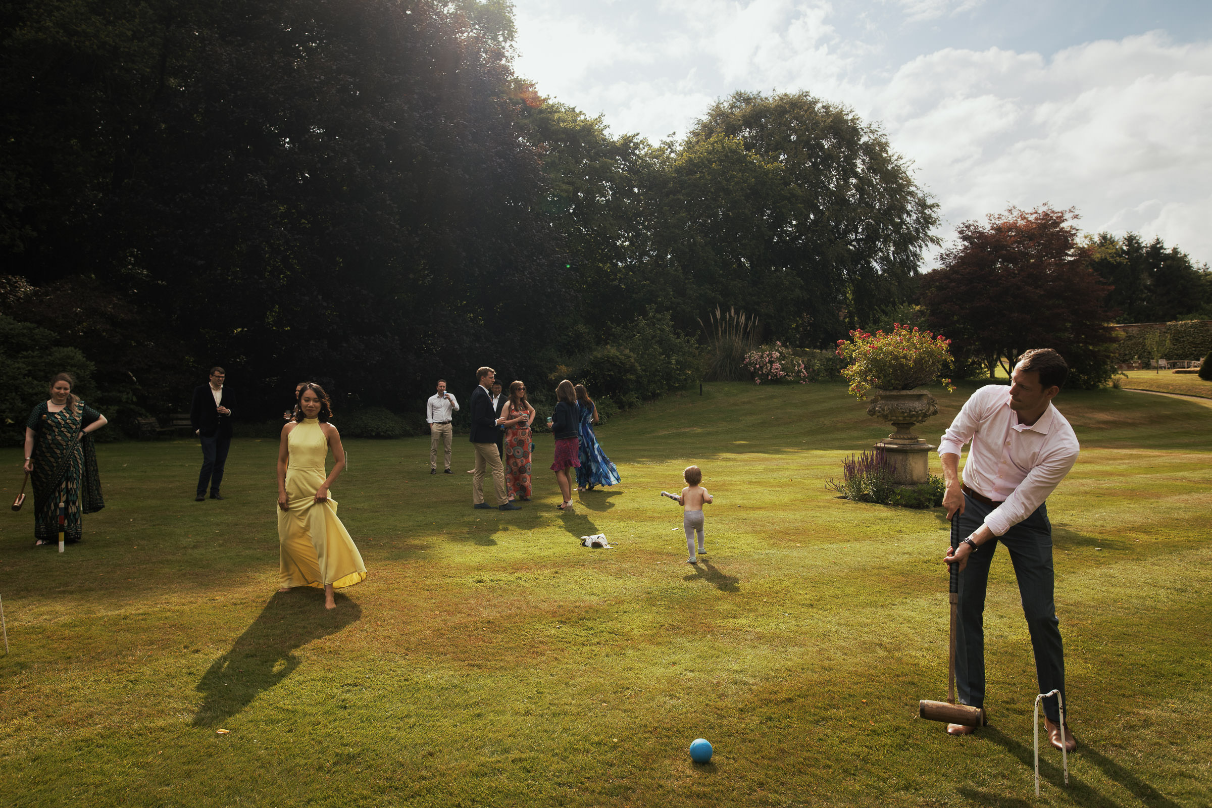 An example of a wedding reception image captured in a documentary style. Multiple moments fill the frame. Man holding a croquet mallet, a barefoot bridesmaid lifting her dress as she walks across the lawn, and a toddler wandering off while wedding guests chat on the main lawn of Homme House.