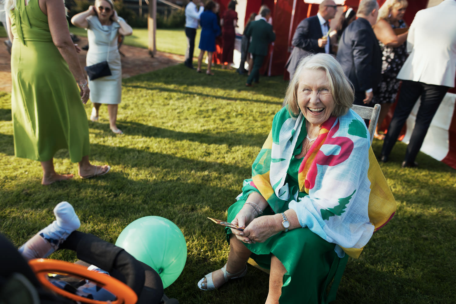 Woman in a green dress and a colourful shawl, sat in a chair and laughing at the camera. Candid moment.