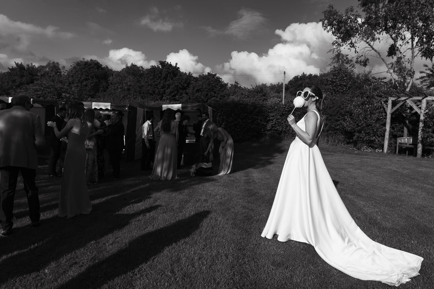 Alpheton Hall Barns Candid Wedding Photography: Bride in a white satin dress and oversized sunglasses enjoys candy floss in the sun. Guests have fun playing fairground games.