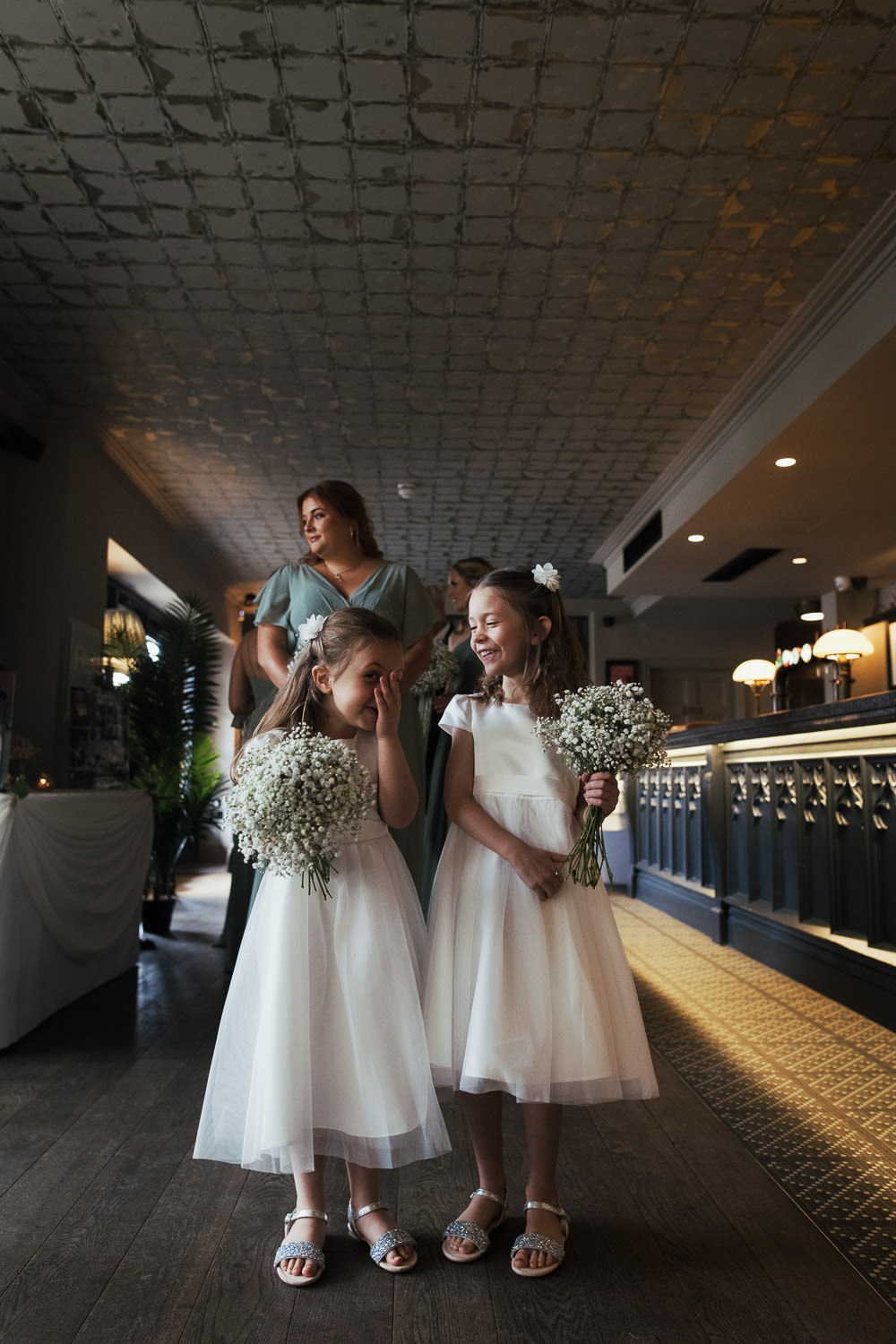 Flower girls in ivory tulle bridesmaid dresses from Monsoon giggle while waiting in the bar for the wedding ceremony at The Lion House, Chelmsford, Essex (CM3 3JA). A bridesmaid in a Mori Lee chiffon dress (style #21667) with flutter sleeves and a front slit stands nearby, creating a joyful and candid wedding moment.