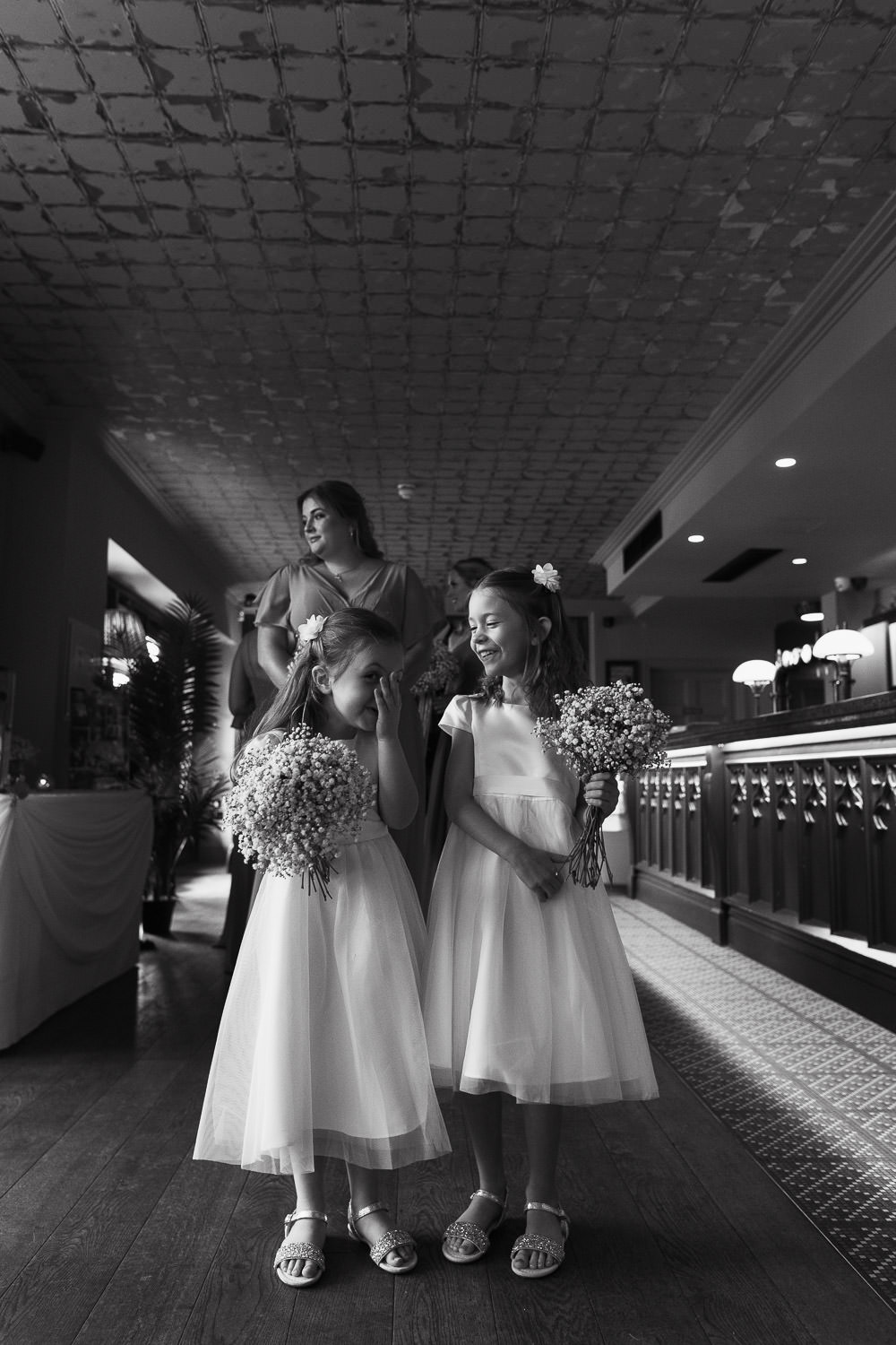Flower girls in ivory tulle bridesmaid dresses from Monsoon giggle while waiting in the bar for the wedding ceremony at The Lion House, Chelmsford, Essex (CM3 3JA). A bridesmaid in a Mori Lee chiffon dress (style #21667) with flutter sleeves and a front slit stands nearby, creating a joyful and candid wedding moment.