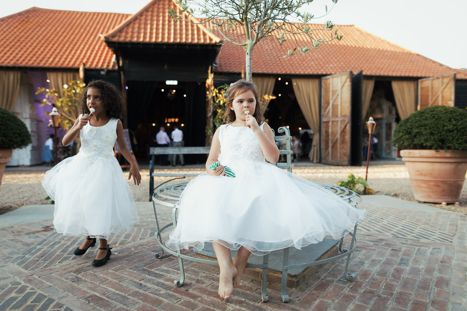 The flower girls are sat outside the Kentish Barn in white dresses, they are eating lollipops.