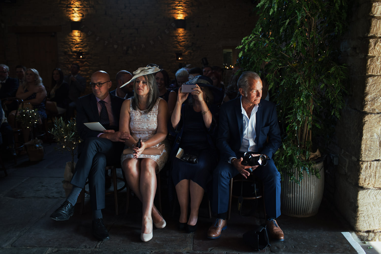 Four guests seated, waiting for a wedding to begin at Cripps Barn. Documentary-style wedding photography. Cotswolds wedding barn GL7 5BA