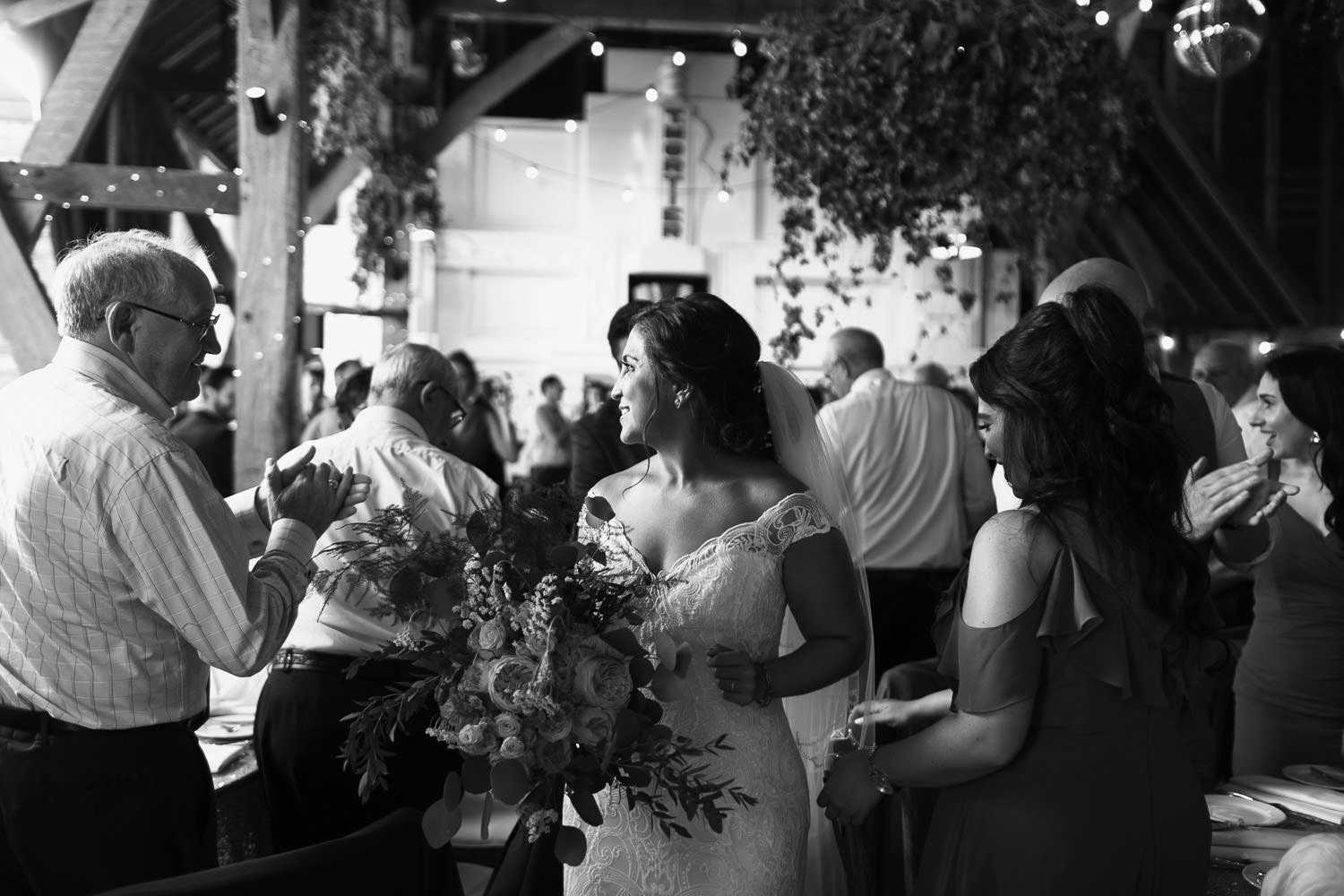 Bride making her way through tables of guests in the Kentish barn as people clap.