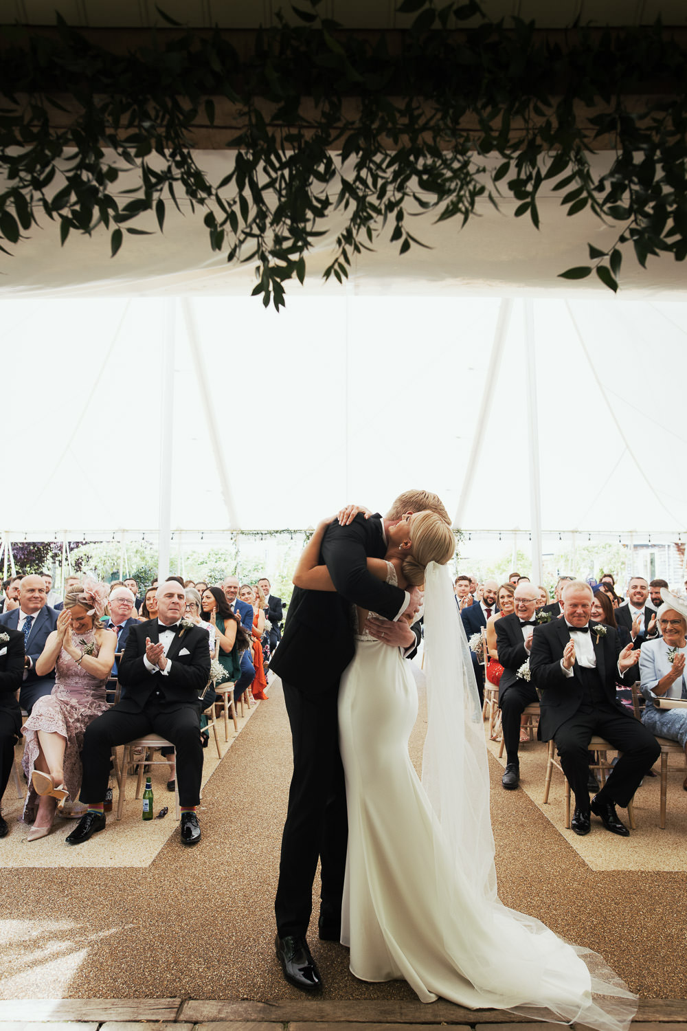 Couple hugging as friends and family clap at the end of their wedding ceremony in the marquee outside at Houchins venue. Groom in suit by The Groom’s Room, bride in ‘Jack’ dress by Made With Love from Halo + Wren Bridal. Captured by top Essex wedding photographer by Tracy Morter.