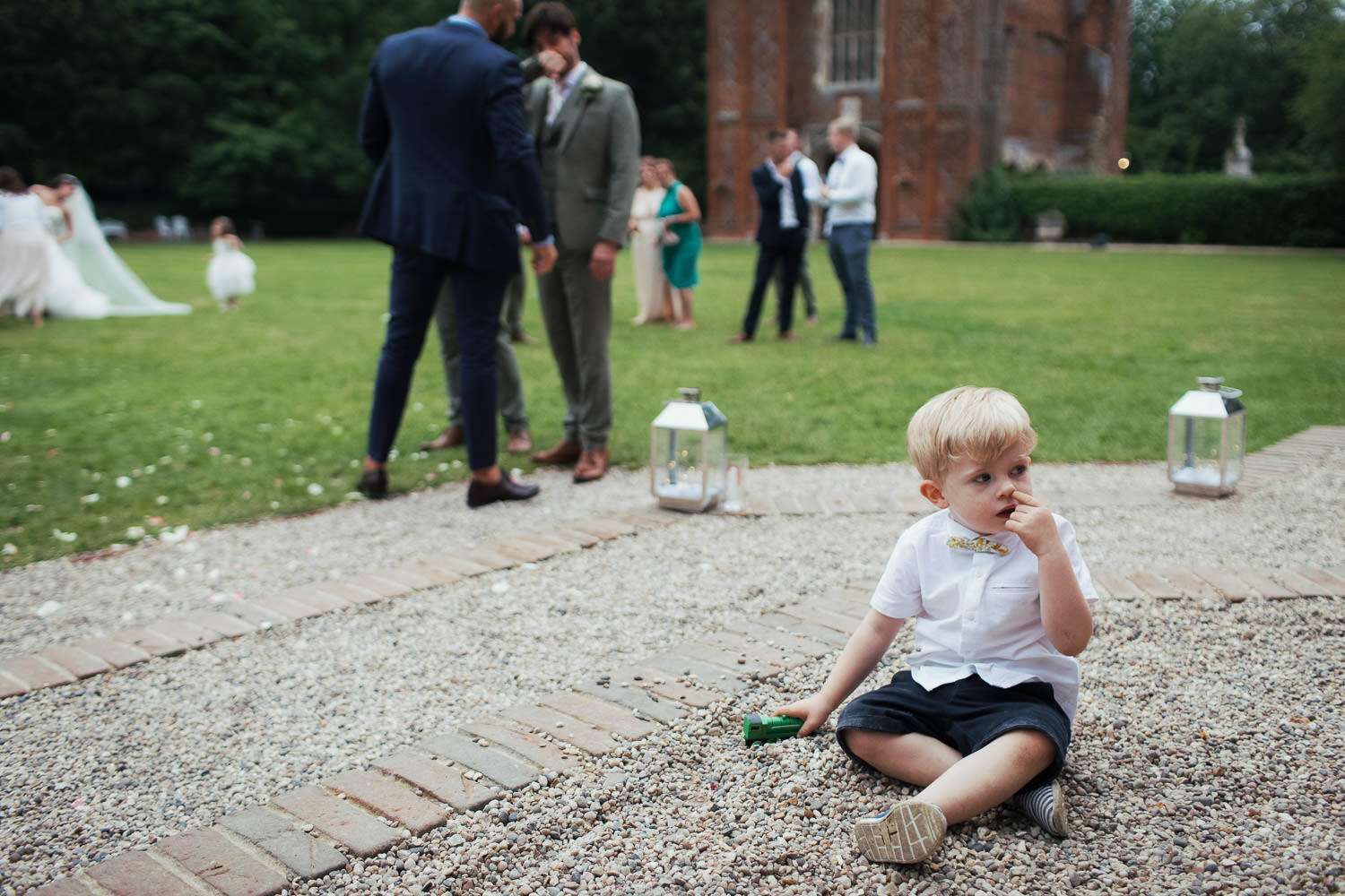 Little boy holding a green Thomas & Friends Henry train toy. Sat on the gravel at a wedding reception while he picks his nose. An historical Tudor Country House Weddings venue in Essex.