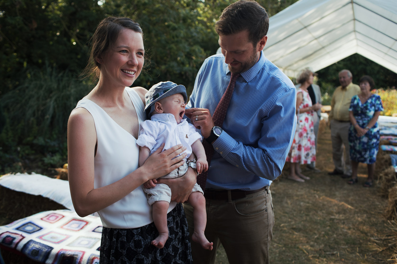 A young baby yawns with his parents at a humanist wedding in a garden.