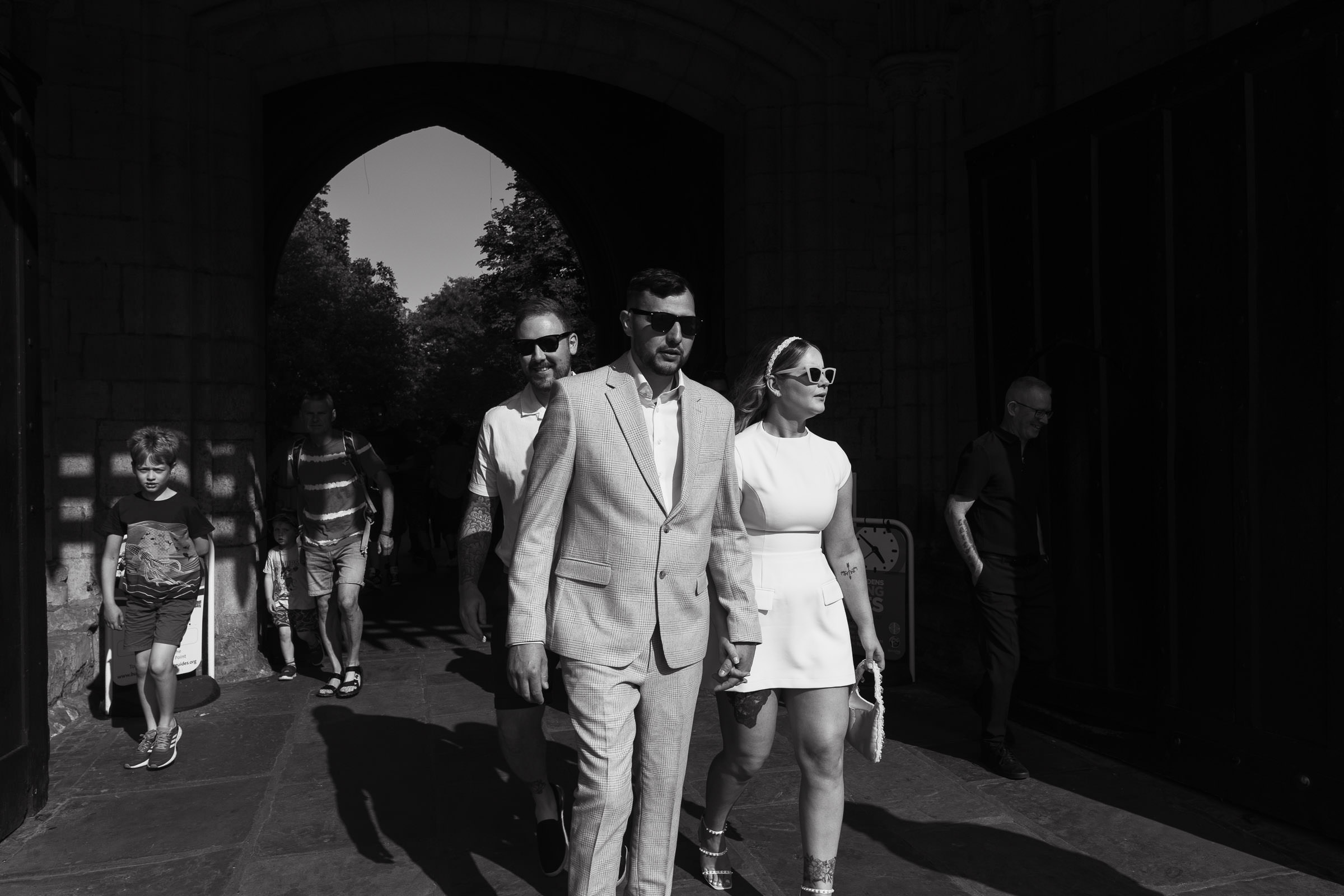 Suffolk Micro-Wedding: Newlyweds holding hands and wearing sunglasses. Walking out of the Abbey Gardens through the Abbey Gate in the sunshine. Bury St Edmunds.