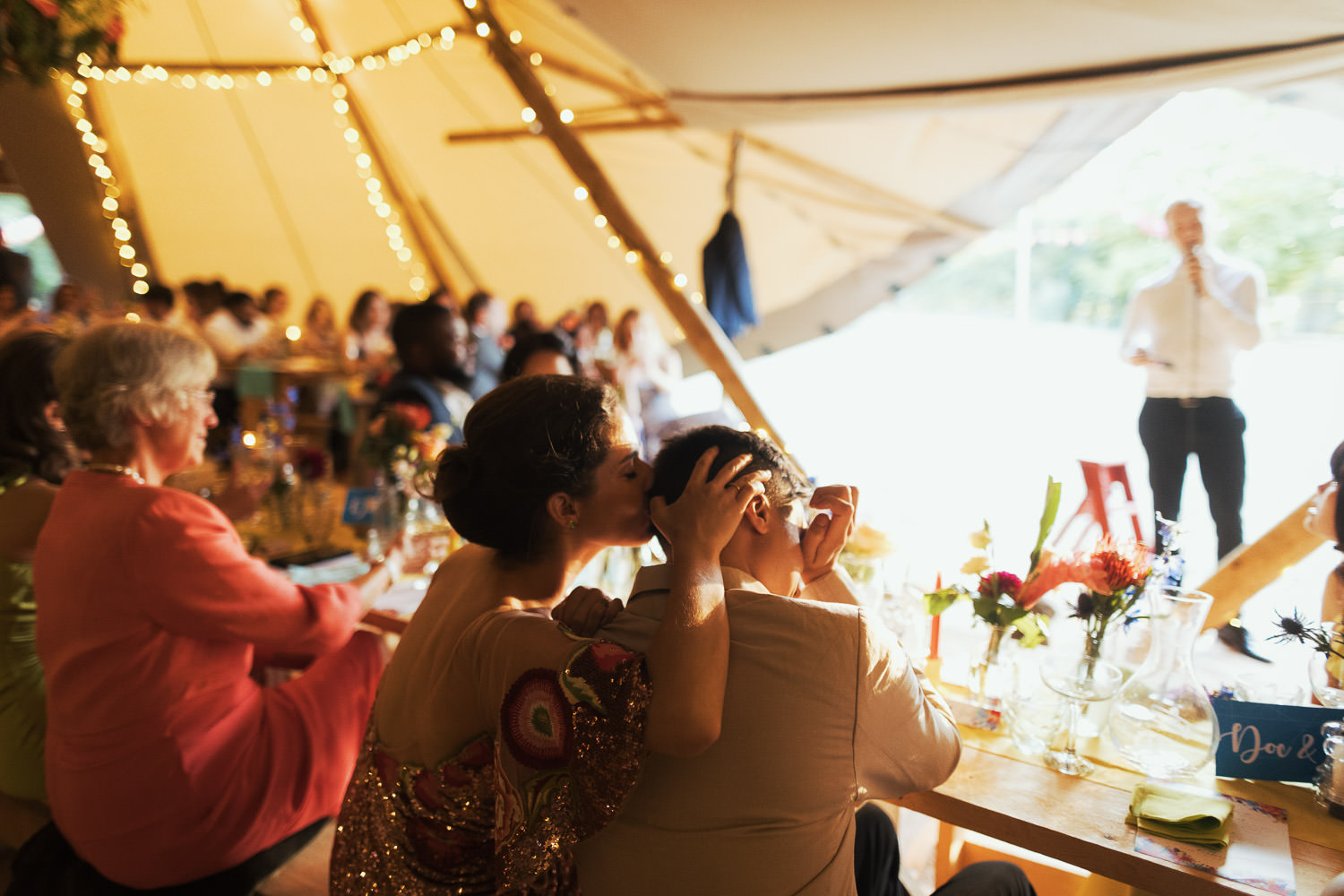 Bride wearing an Alice Temperley Carnation sequin gown, kissing her husband on his head during emotional wedding speeches. Candid documentary photography.