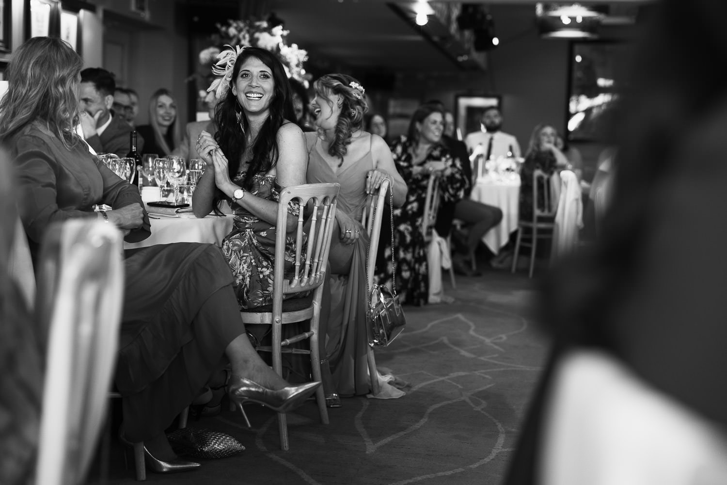 Woman with long dark hair and huge smile, holds her hands together. She's wearing a FLORAL PRINT SATIN DRAPED BARDOT MIDI DRESS from Pretty Little Thing. She's watching the speeches at a wedding reception at The Lion House.