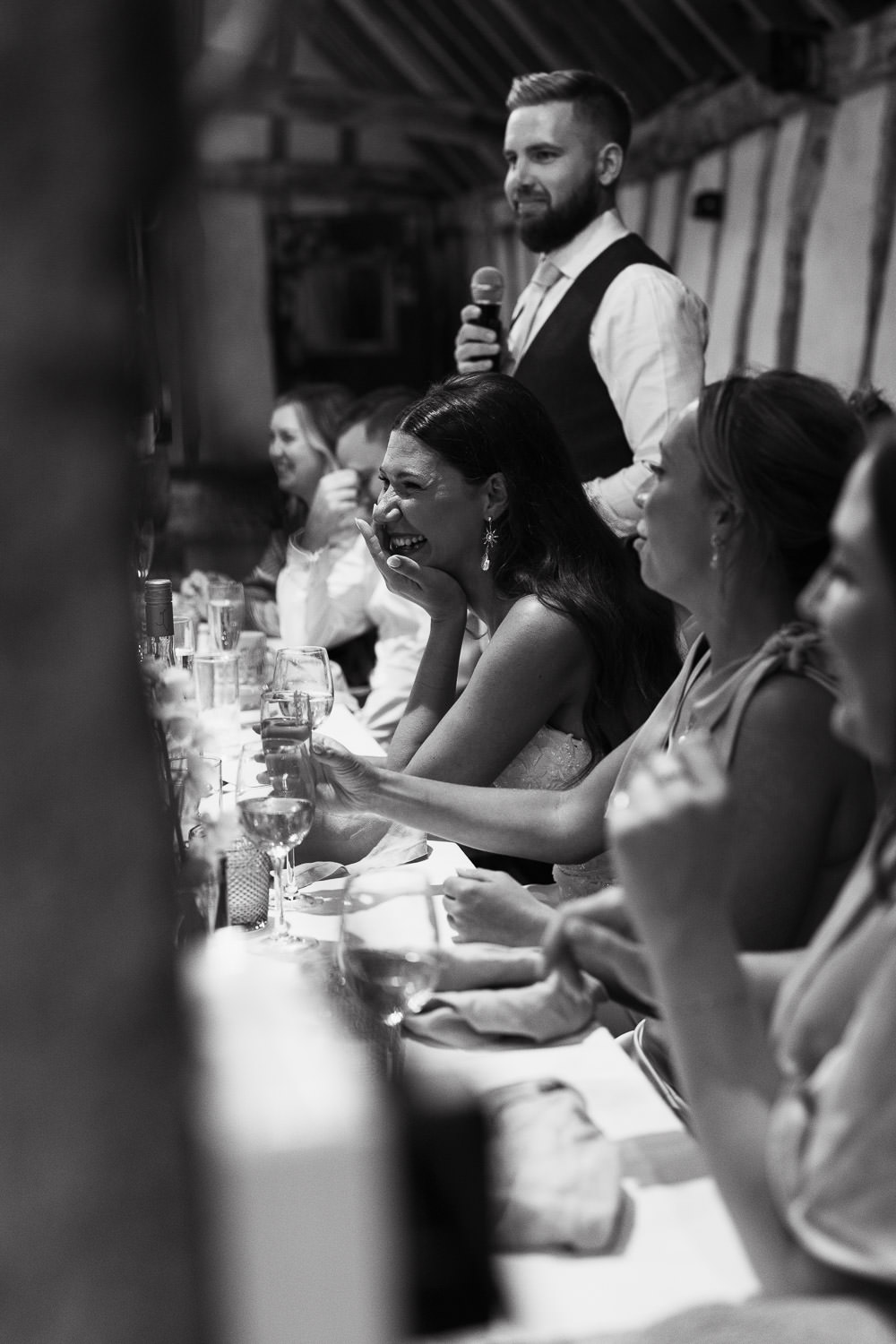 Alpheton Hall Barns Wedding Photographer: Bride covers her mouth as she laughs during her new husband's speech. At the top table.