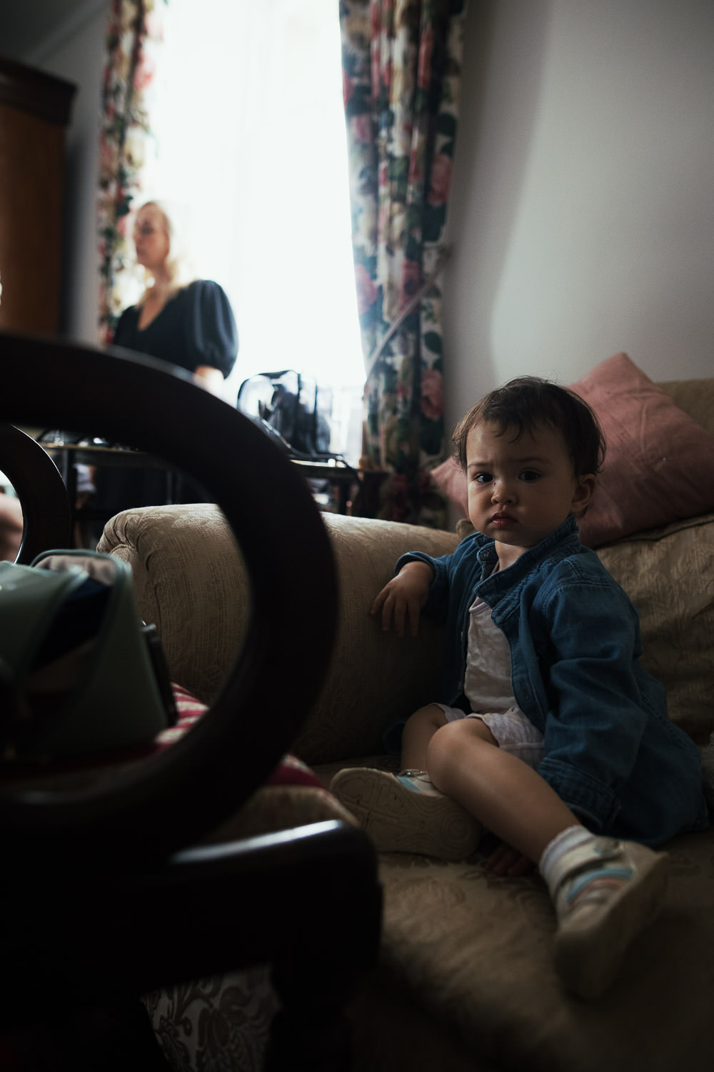 A toddler wearing trainers and a denim jacket sits on a chair and looks at the camera.Waiting while people get ready for wedding. The makeup artist in the background by the window.