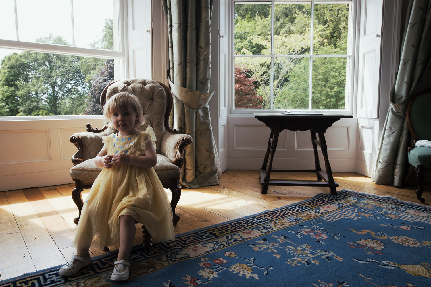 A little girl in a yellow dress is sitting on a stool in the bedroom of a wedding venue called Homme House. There are windows and trees in the background.JoJo Maman Bébé Floral Yellow Floral Tulle Party Dress.