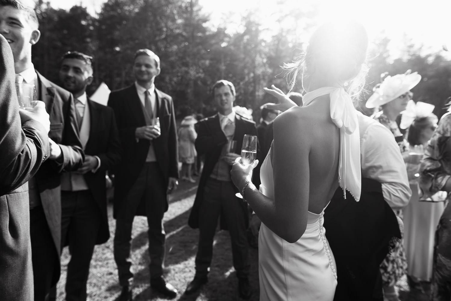 View from behind a bride on her wedding day, drinks in the sun. She's holding glass of champagne, men in the background wearing suits. Candid Norfolk Wedding photography. Katrine Mogensen bespoke bridal gown.